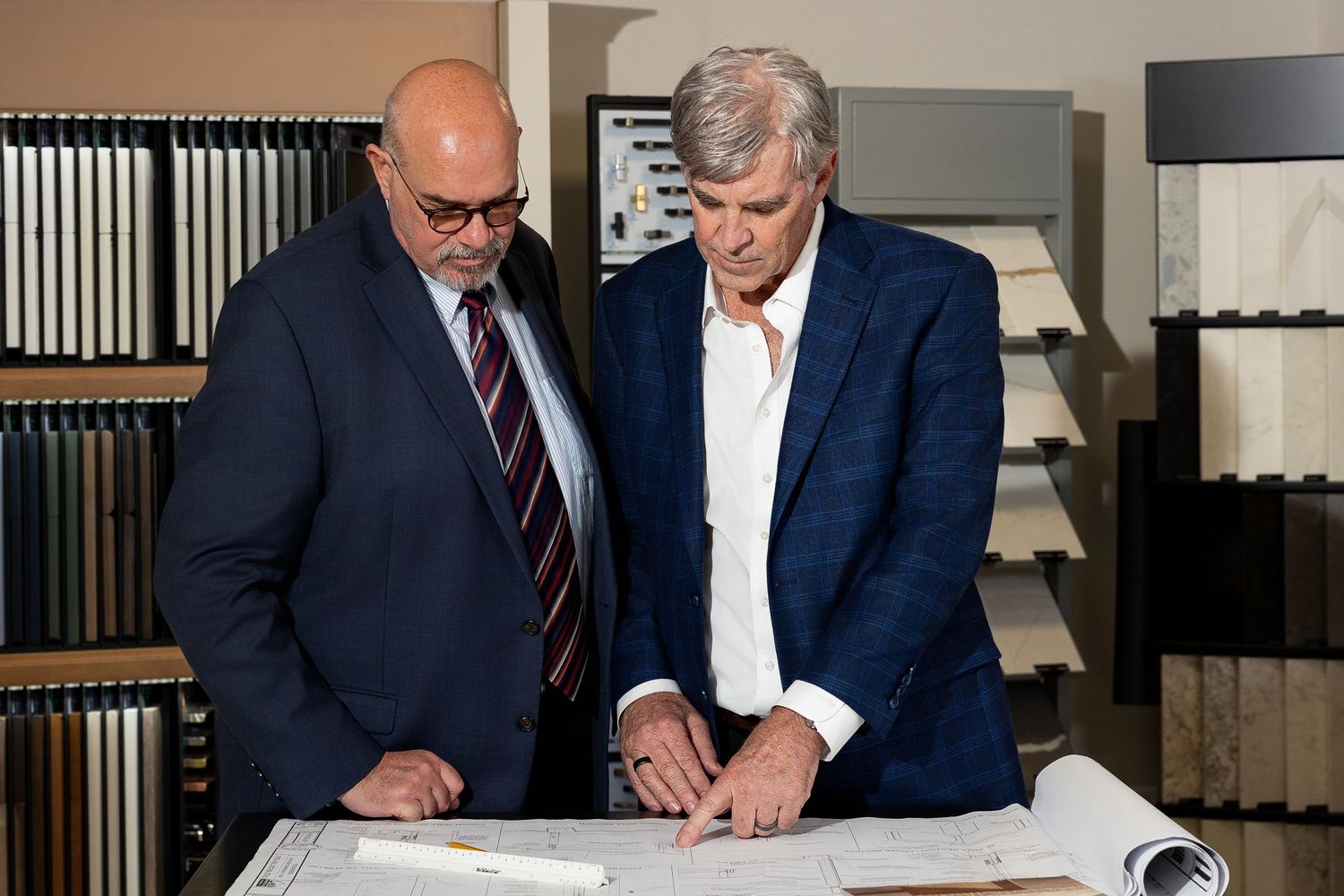 Two men in suits reviewing large architectural blueprints spread on a table in a showroom.