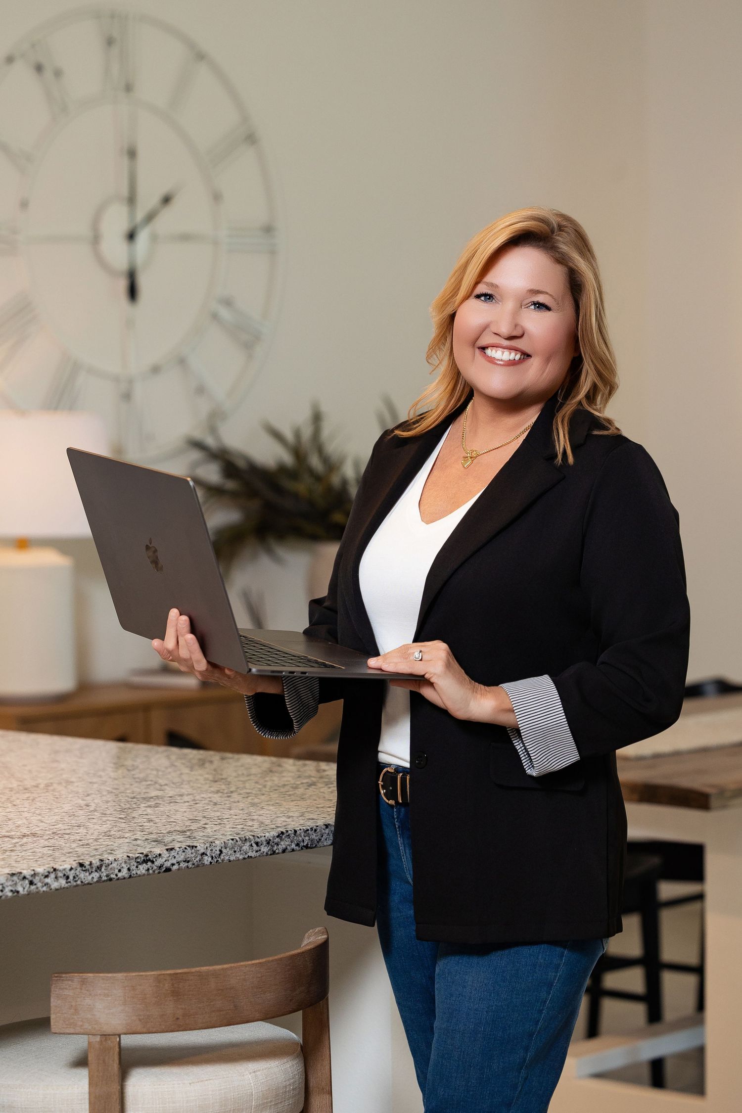 Smiling blonde woman in black blazer holding laptop computer in stylish home kitchen setting.