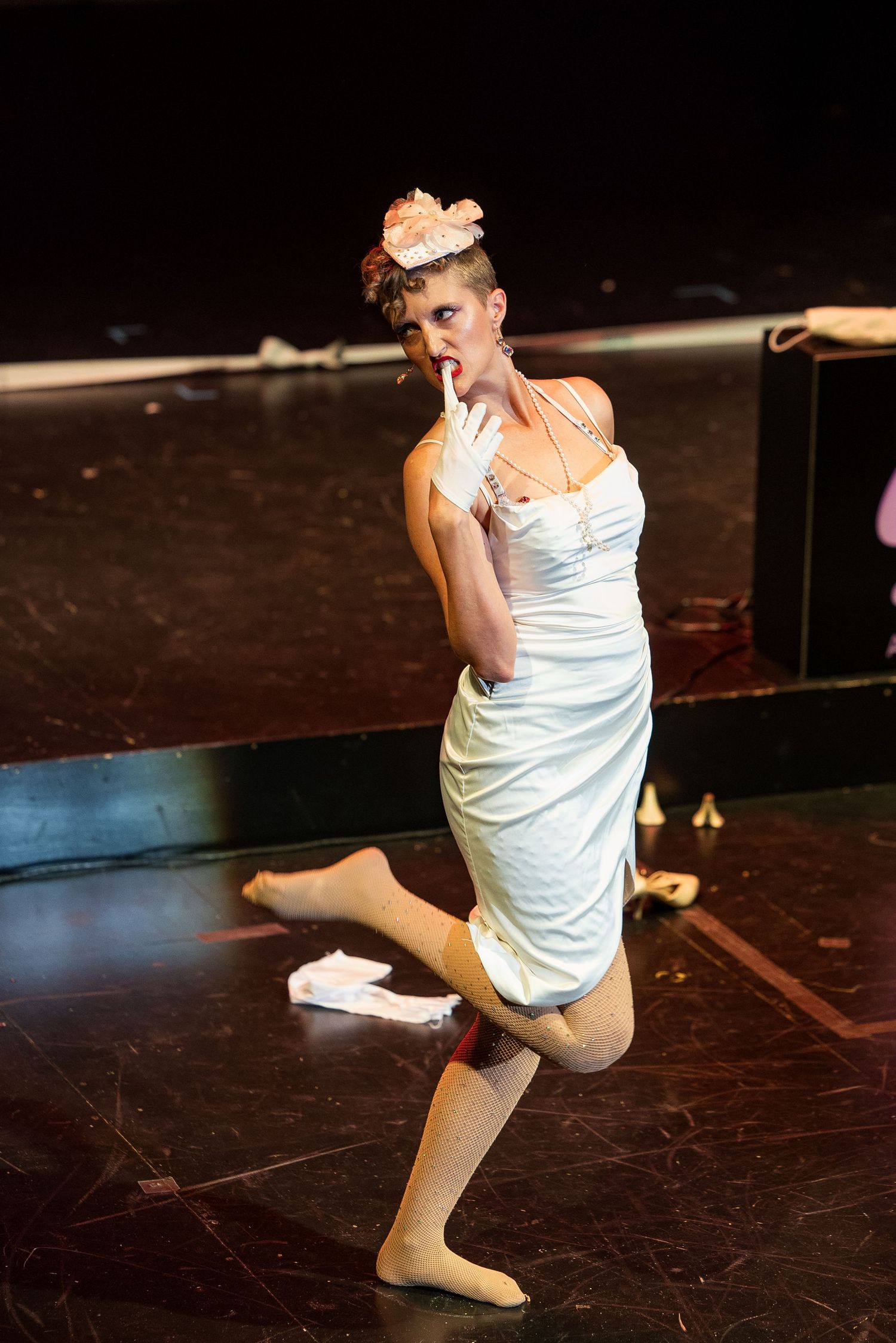 Burlesque performer in white dress with floral hat strikes playful pose with gloved finger to lips.