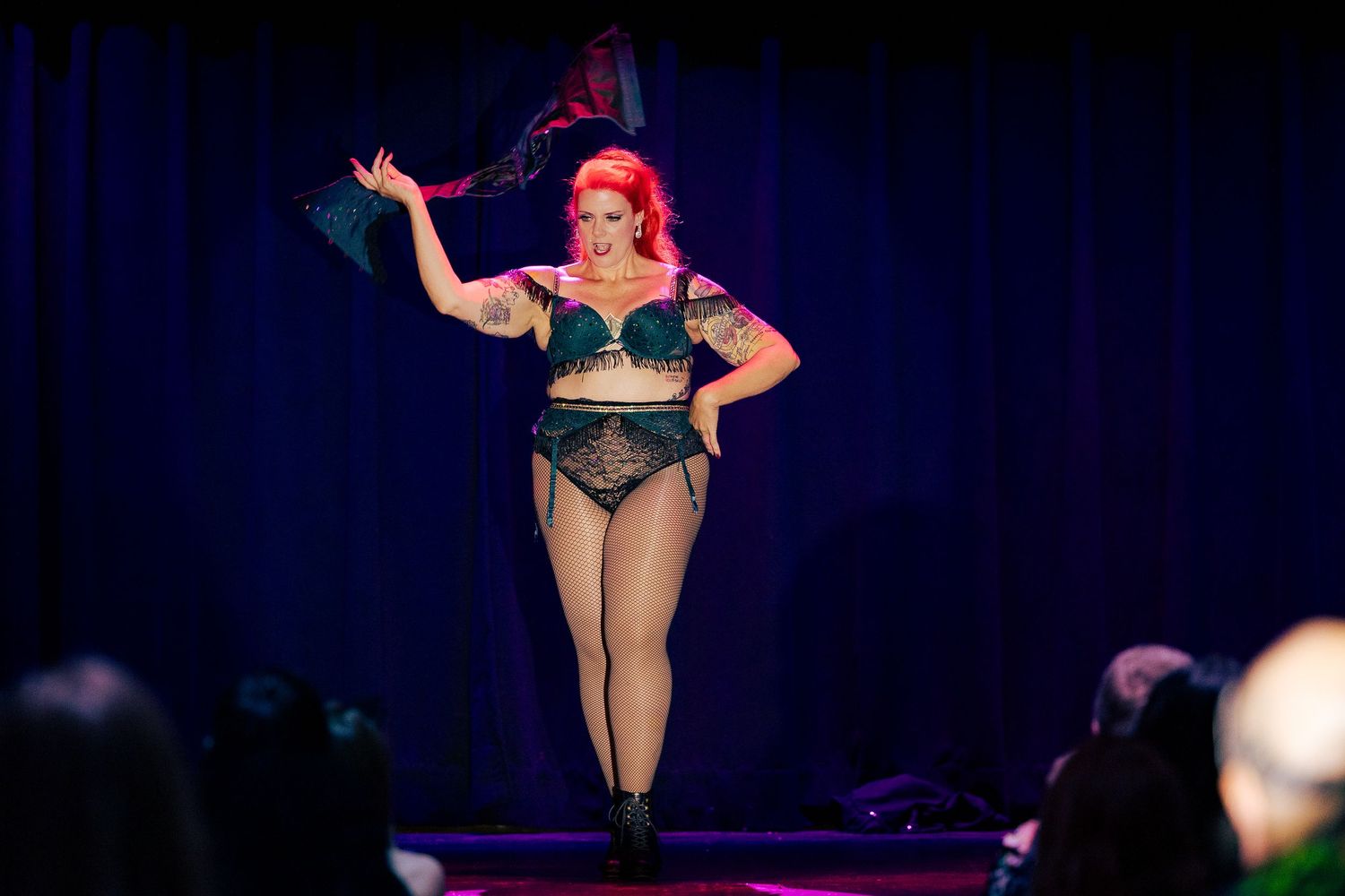 A tattooed redhead burlesque performer in a green lace bra and black stockings dances on stage.