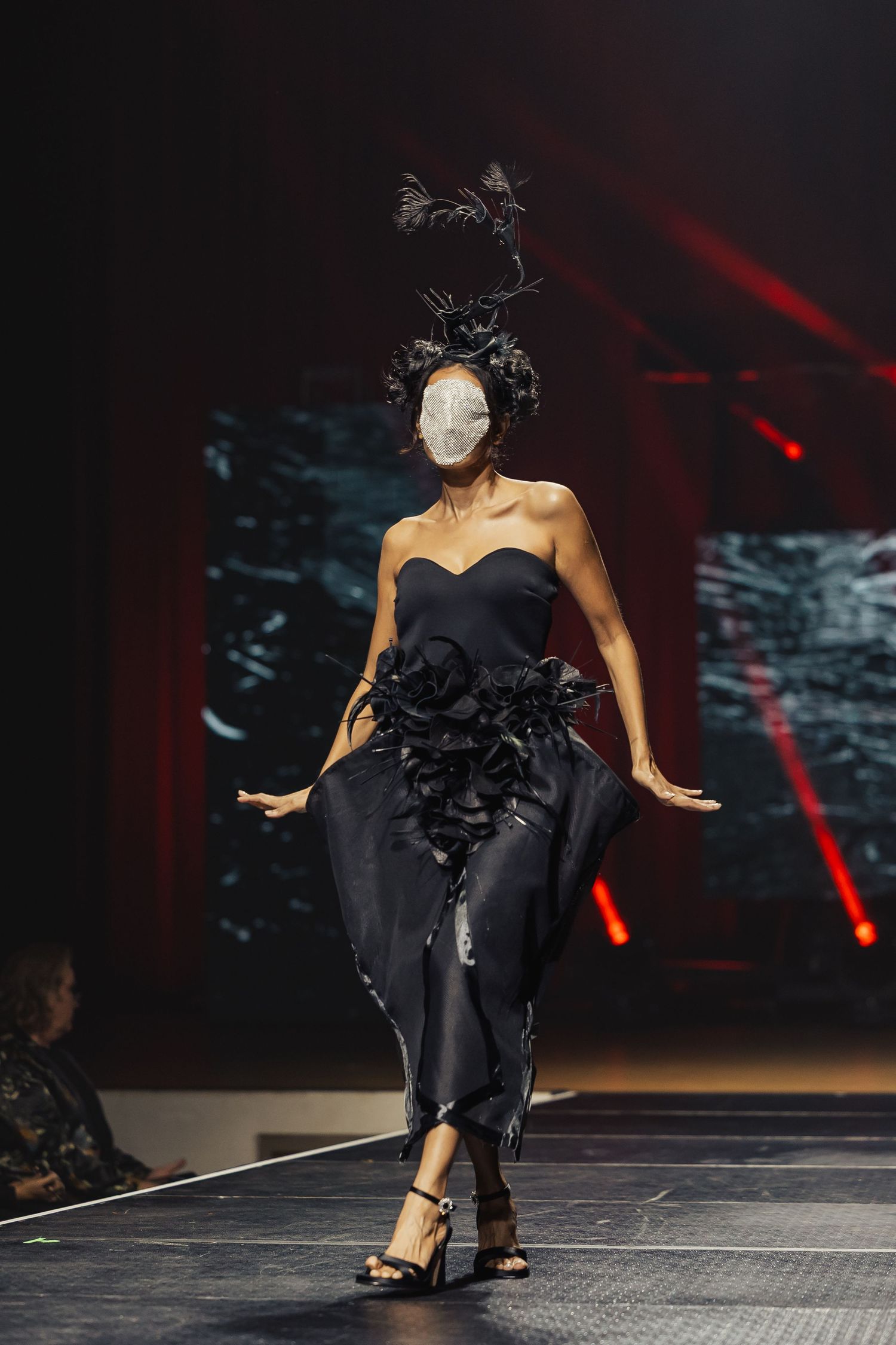 Model in black strapless gown with floral embellishments and feathered headpiece walks fashion runway.
