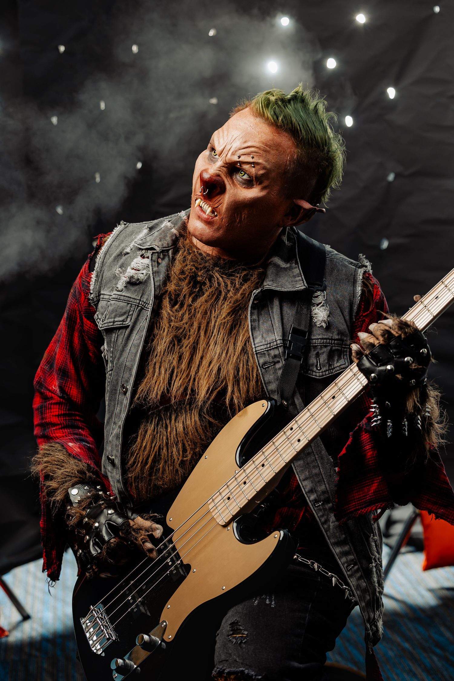 Intense werewolf cosplayer with green mohawk playing bass guitar on smoky stage under spotlights.