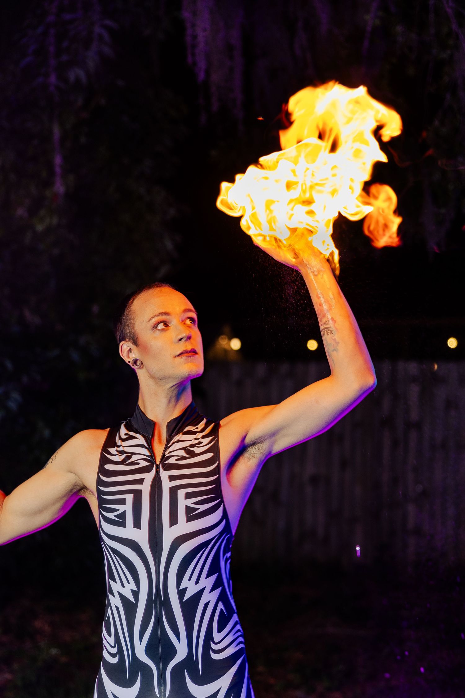 Circus performer in black and white tribal costume holds a large flaming torch aloft at a night event.