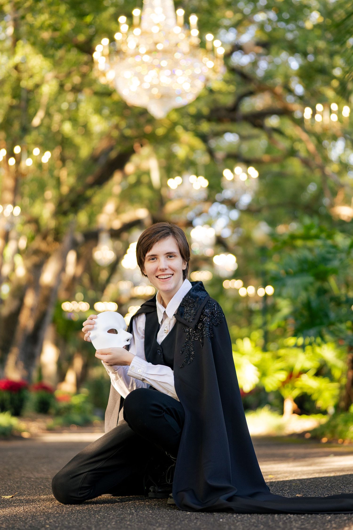 Person in black cape holding white mask kneeling outdoors under chandelier lights among lush green trees.