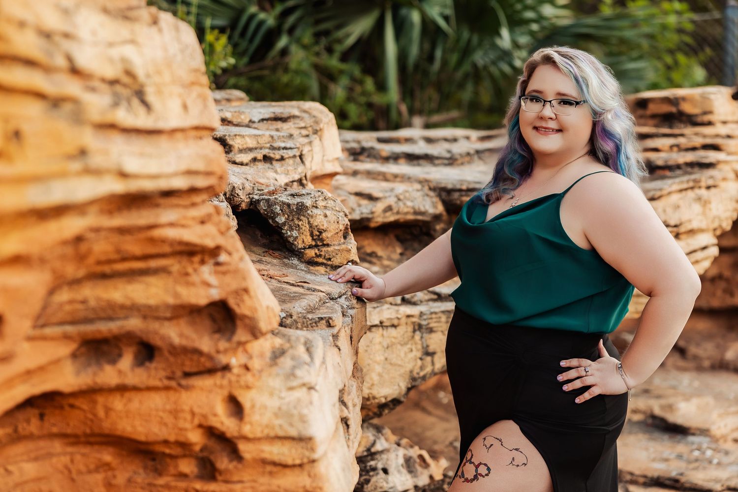 Plus-size woman with purple hair and glasses poses confidently by sandstone rocks in a green top and black skirt.