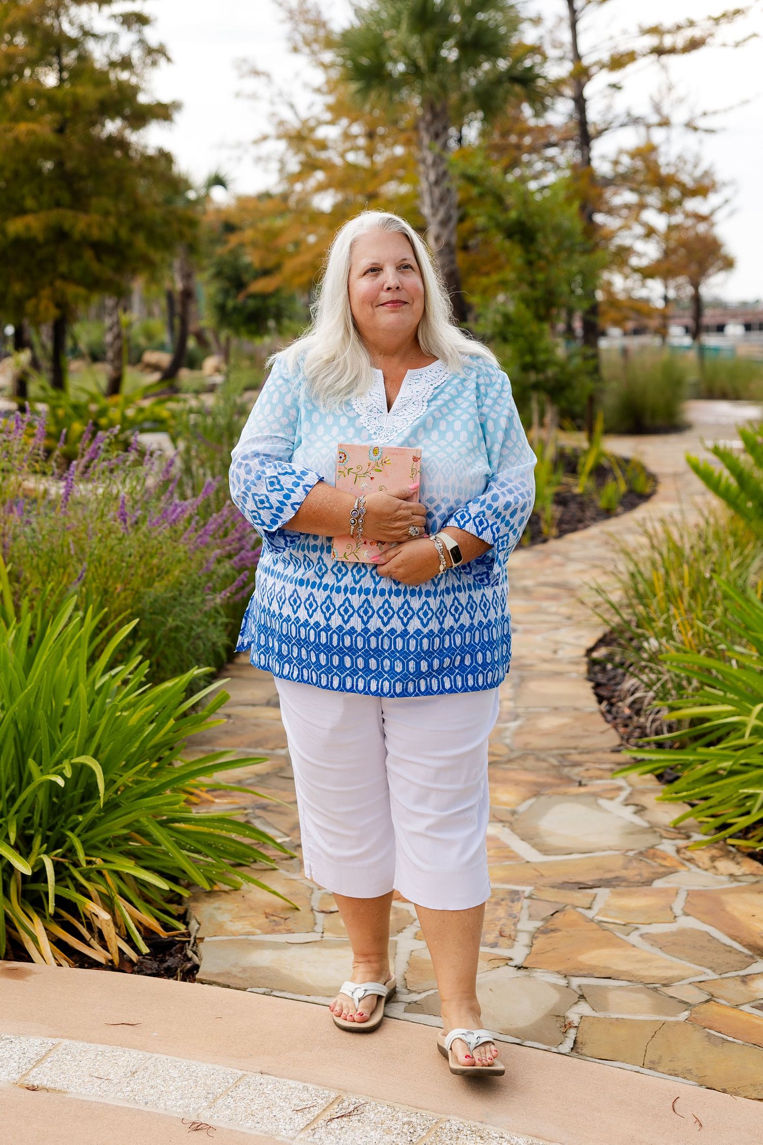 Older woman with white hair wearing blue patterned top and white capris walks along stone garden path holding a book.