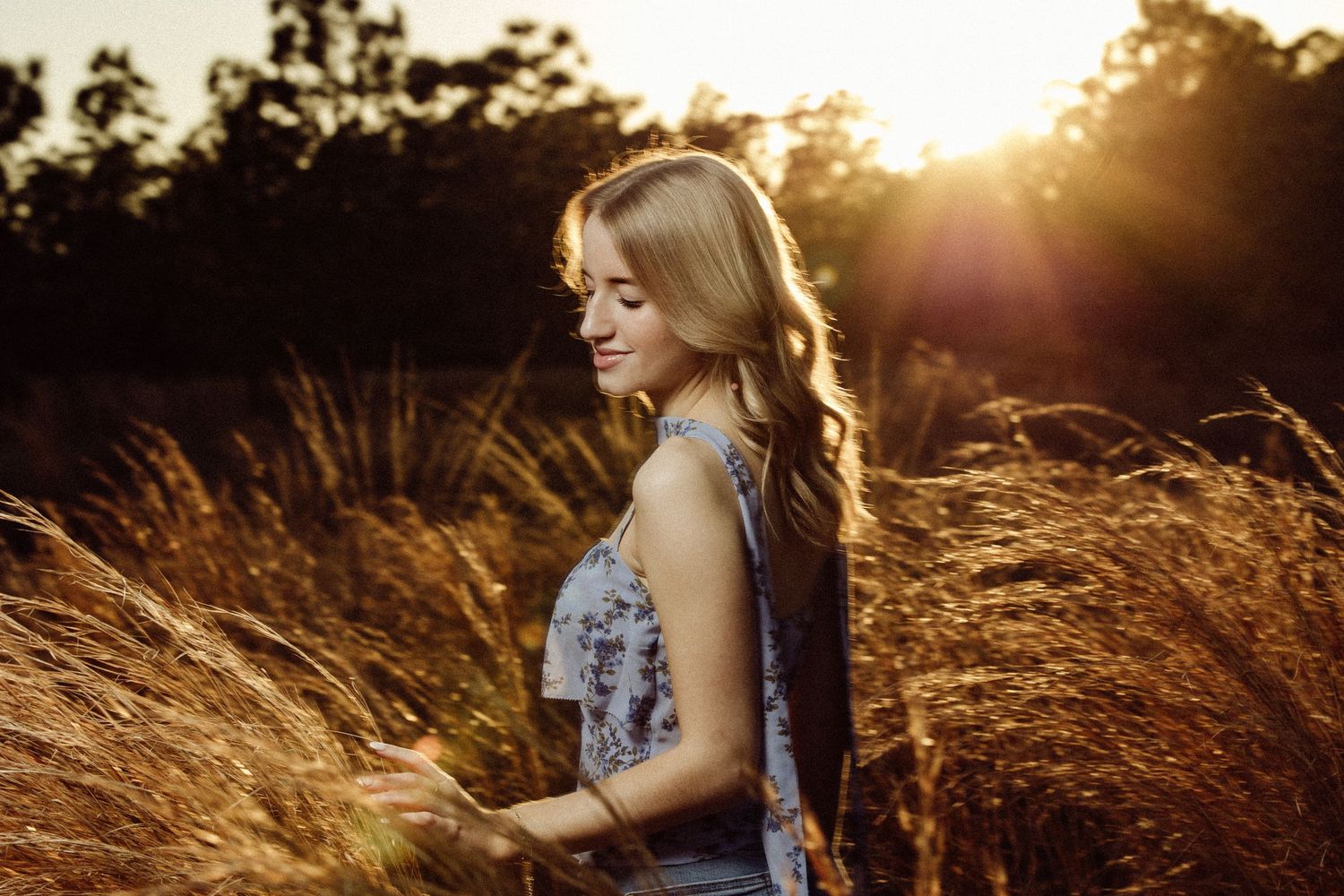 Young woman with long hair smiling in tall grass field during golden hour sunset.
