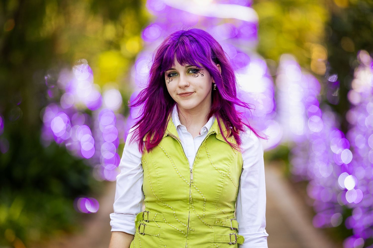Young woman with vibrant purple hair in green vest cosplay poses outdoors with purple bokeh lights.