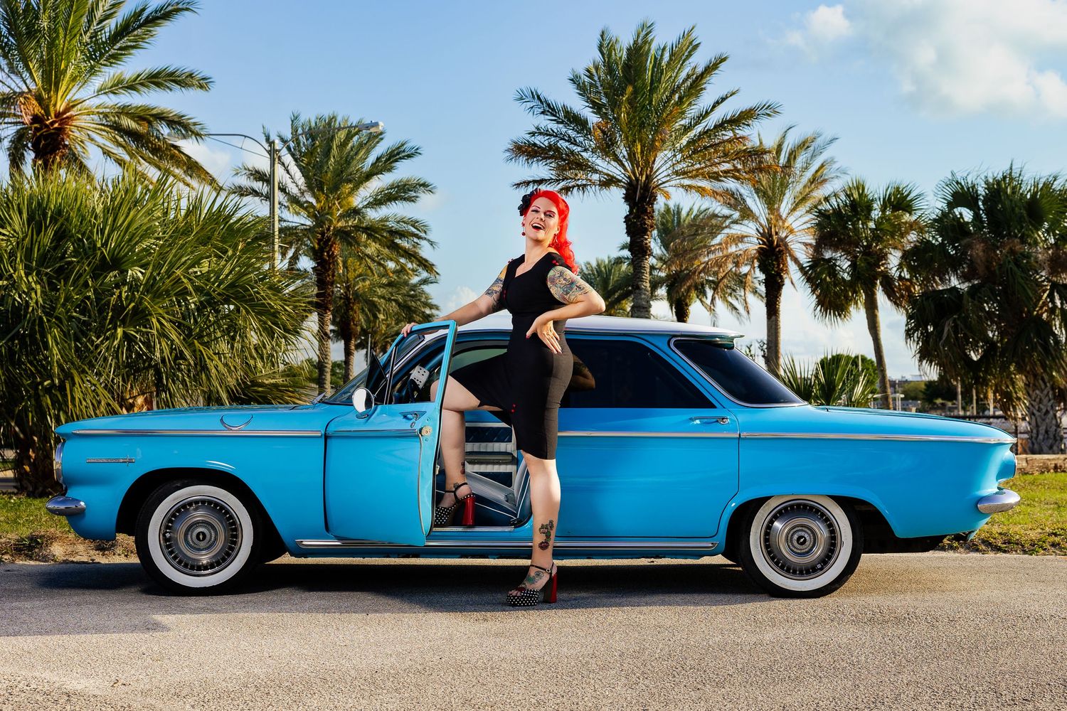 Tattooed redhead woman in black dress stands beside classic blue vintage Corvair among palm trees.