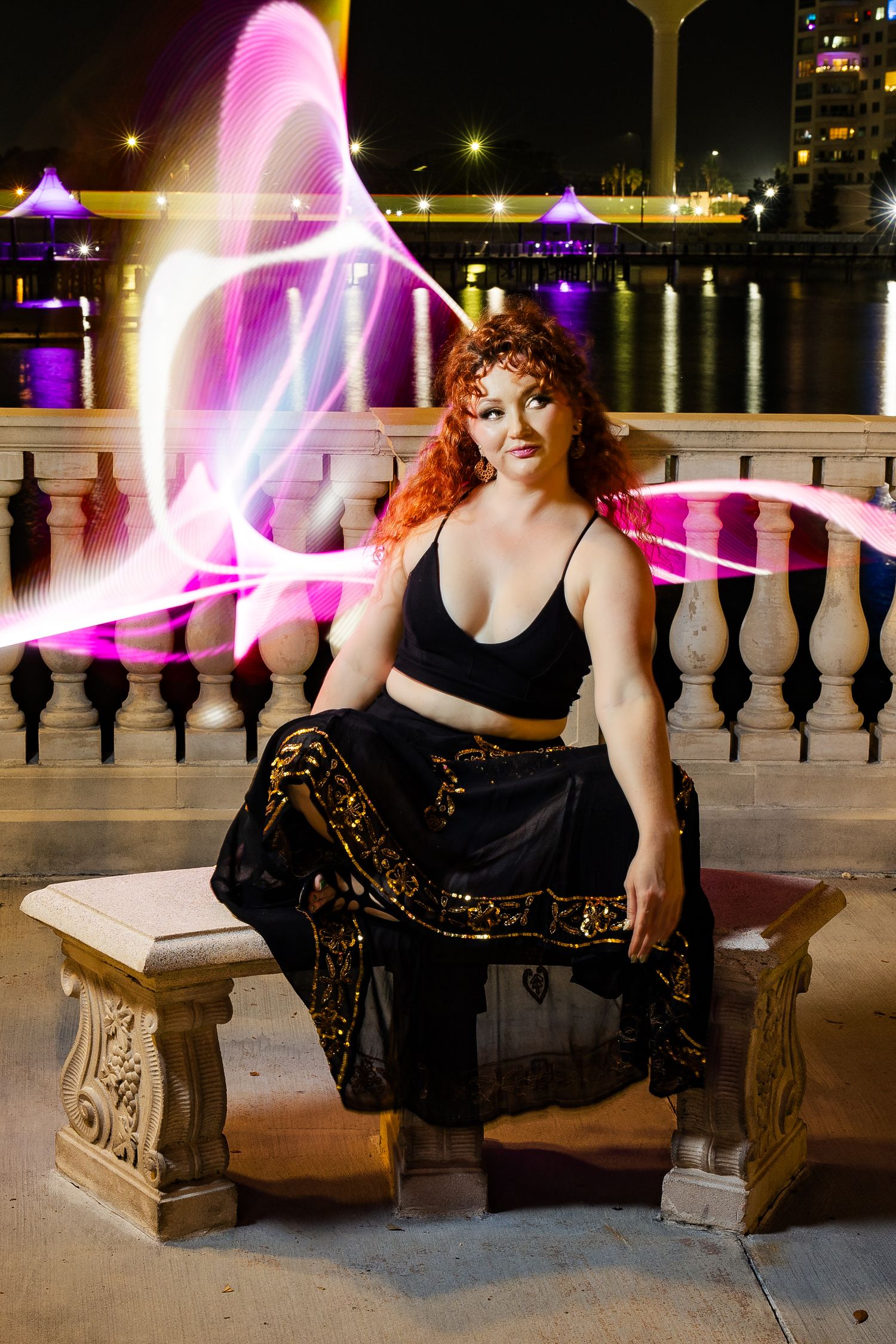 Red-haired woman in black outfit poses on stone bench with pink light painting swirls at night by a waterfront.