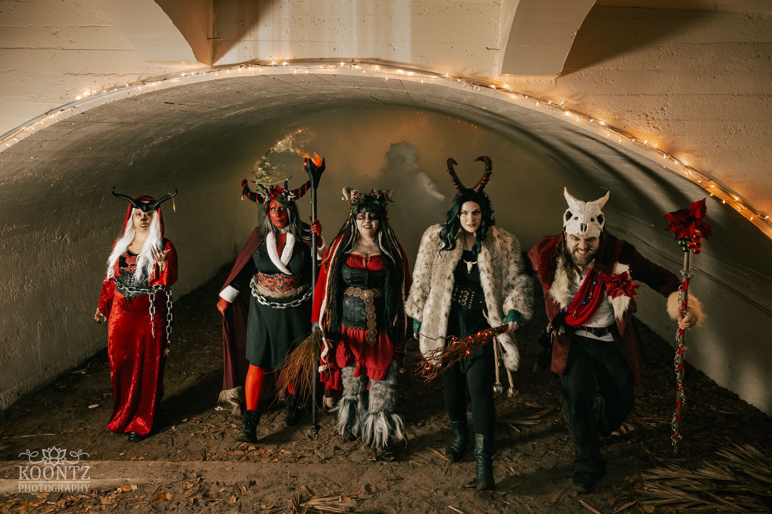 Five cosplayers in detailed fantasy devil and creature costumes pose inside a stone tunnel archway.