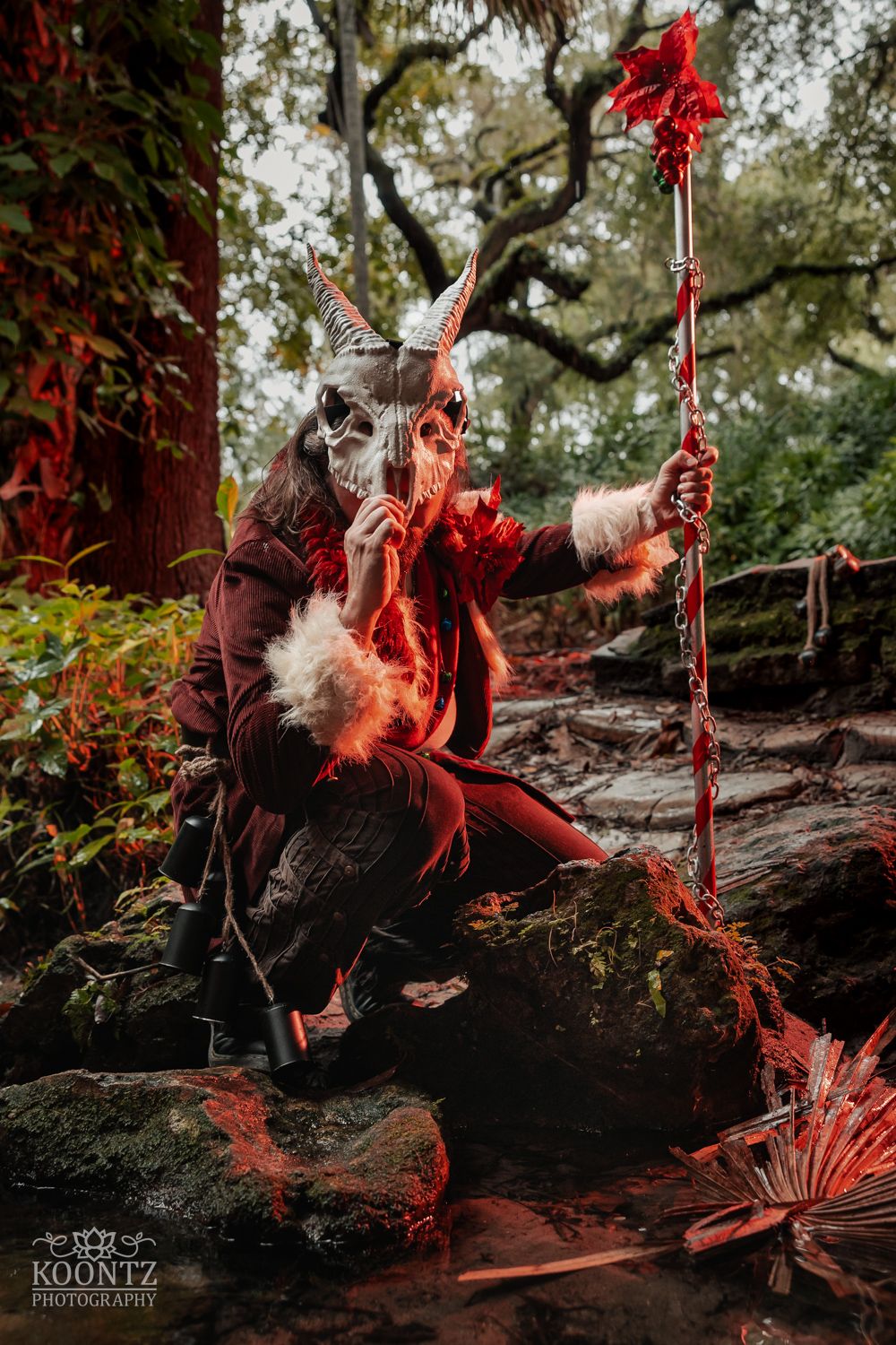 A cosplayer wearing a horned skull mask and red coat crouches on rocks wielding a red floral staff in a forest.
