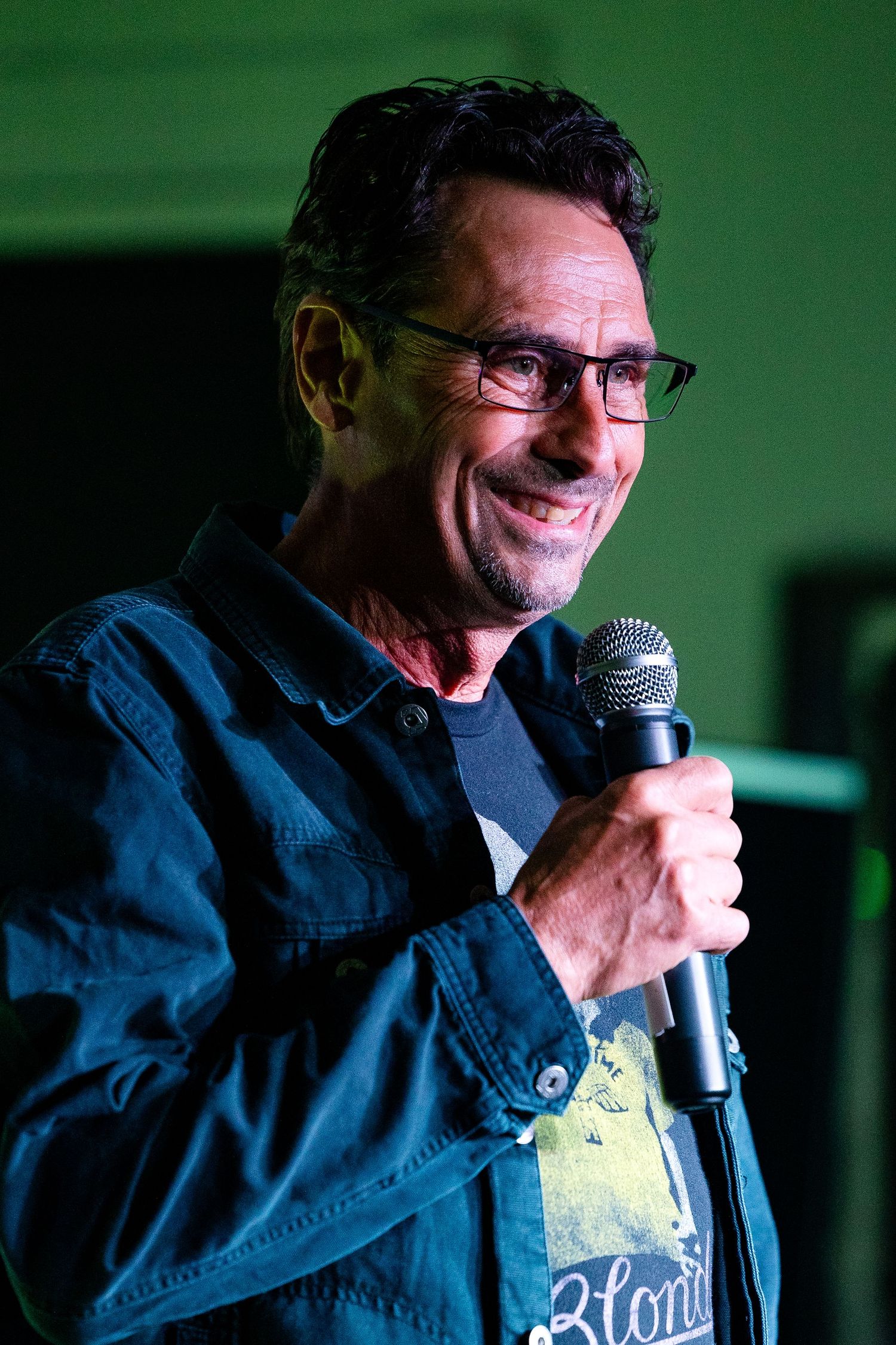 Middle-aged man with glasses smiling while holding a microphone performing stand-up comedy on a dimly lit stage.