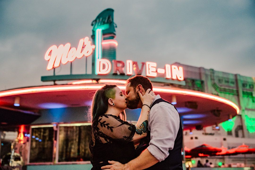 Couple sharing a passionate kiss in front of the iconic Mel's Drive-In neon sign at night.