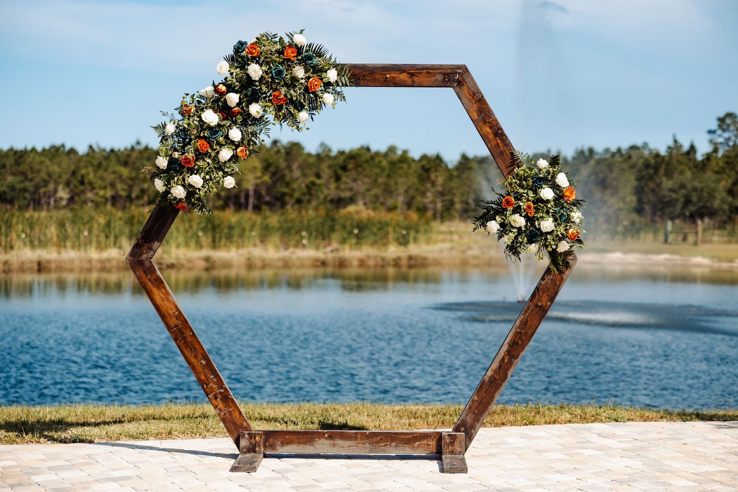 Hexagonal wooden wedding arch decorated with orange and white flowers against a lakeside backdrop.