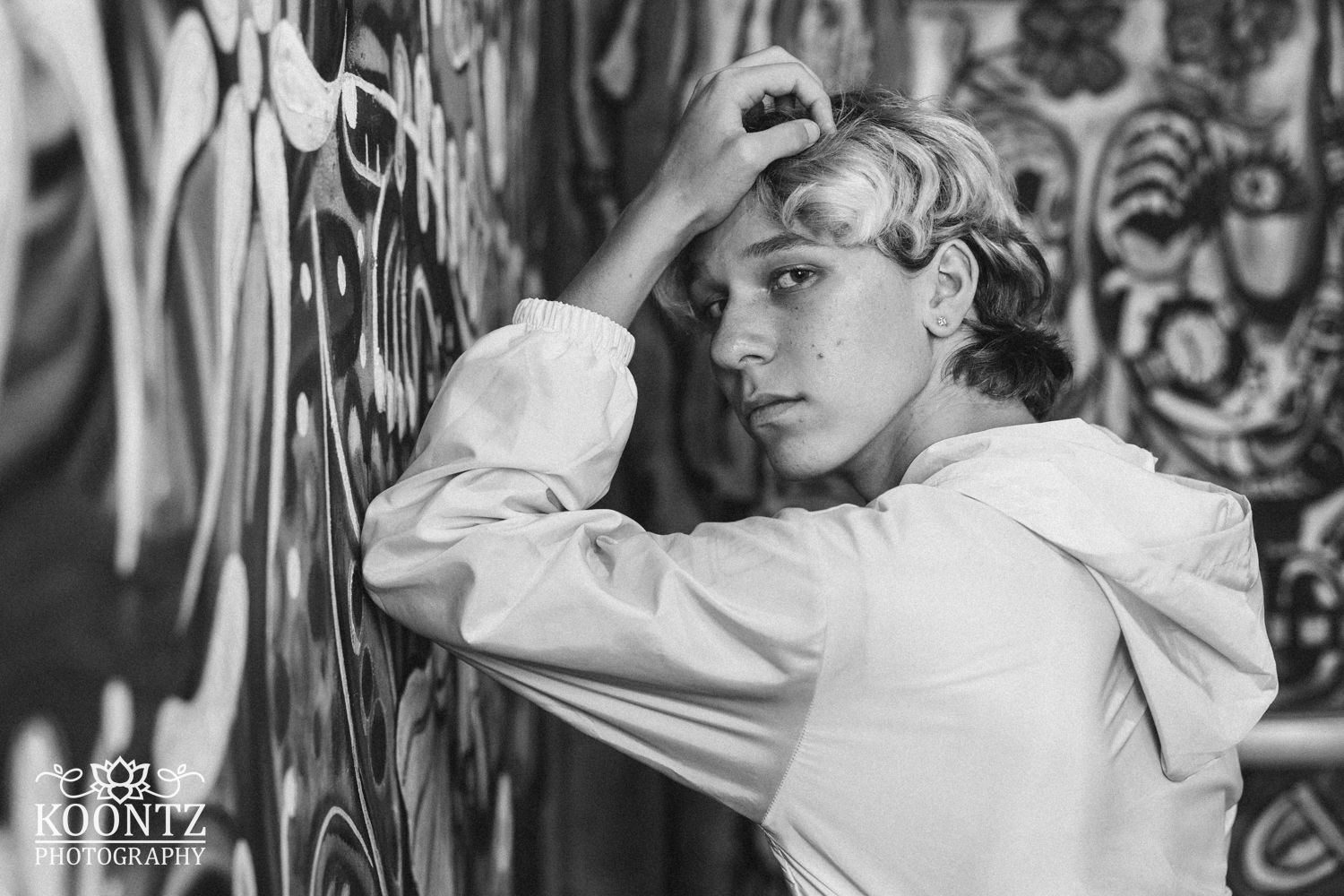 Black and white portrait of a person in white hoodie leaning against graffiti wall.
