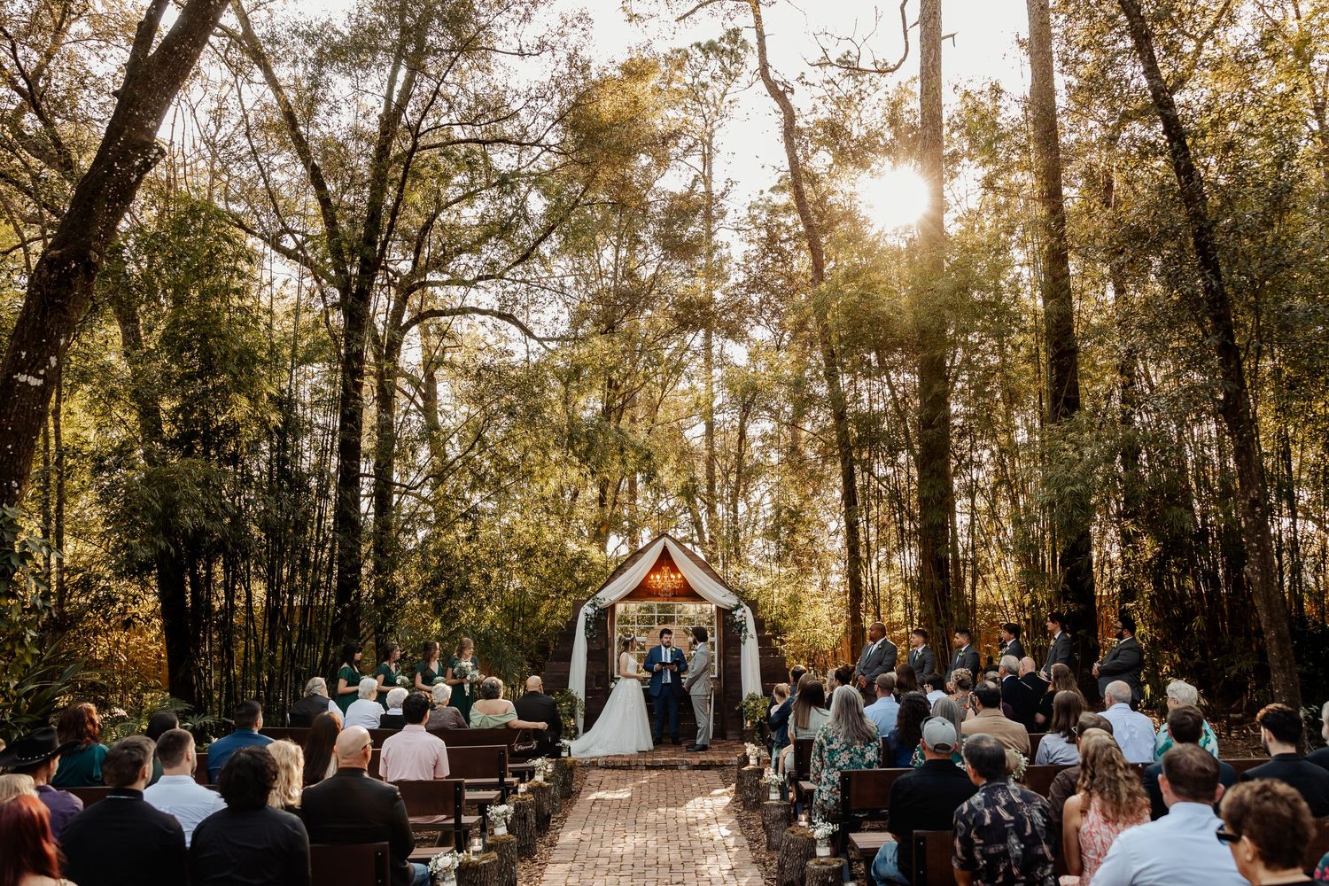 Wide shot of outdoor wedding ceremony in a forest with wooden chapel and sunlight peeking through trees.
