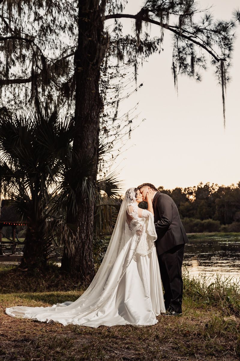 Bride in lace wedding gown and groom kissing near a lake under Spanish moss trees at sunset.
