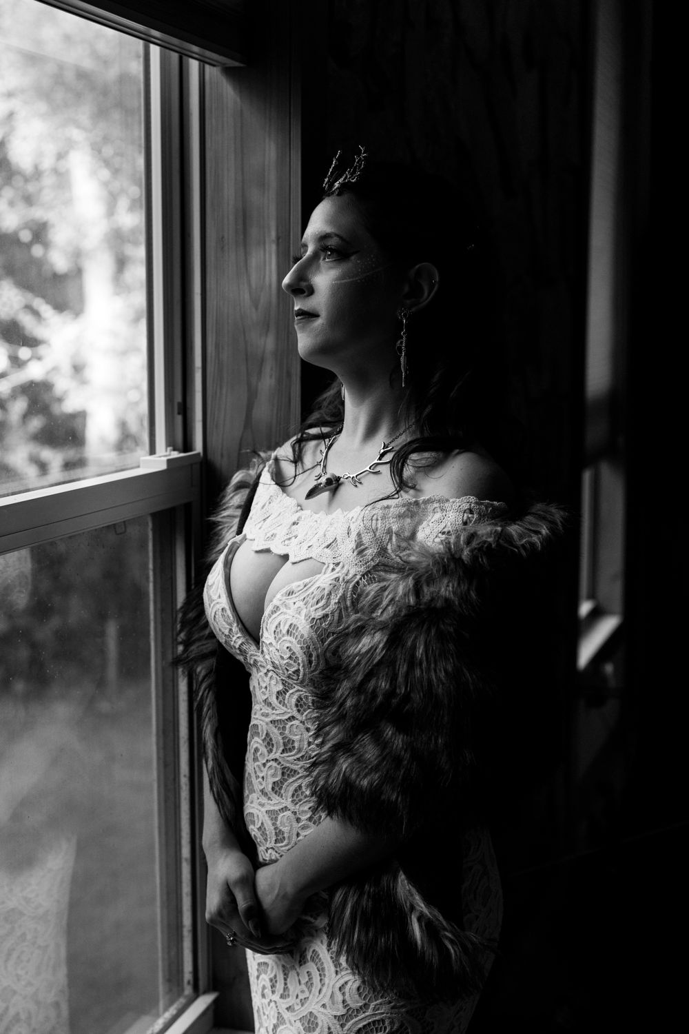 Elegant bride in lace gown and fur stole gazes out window in dramatic black and white portrait.