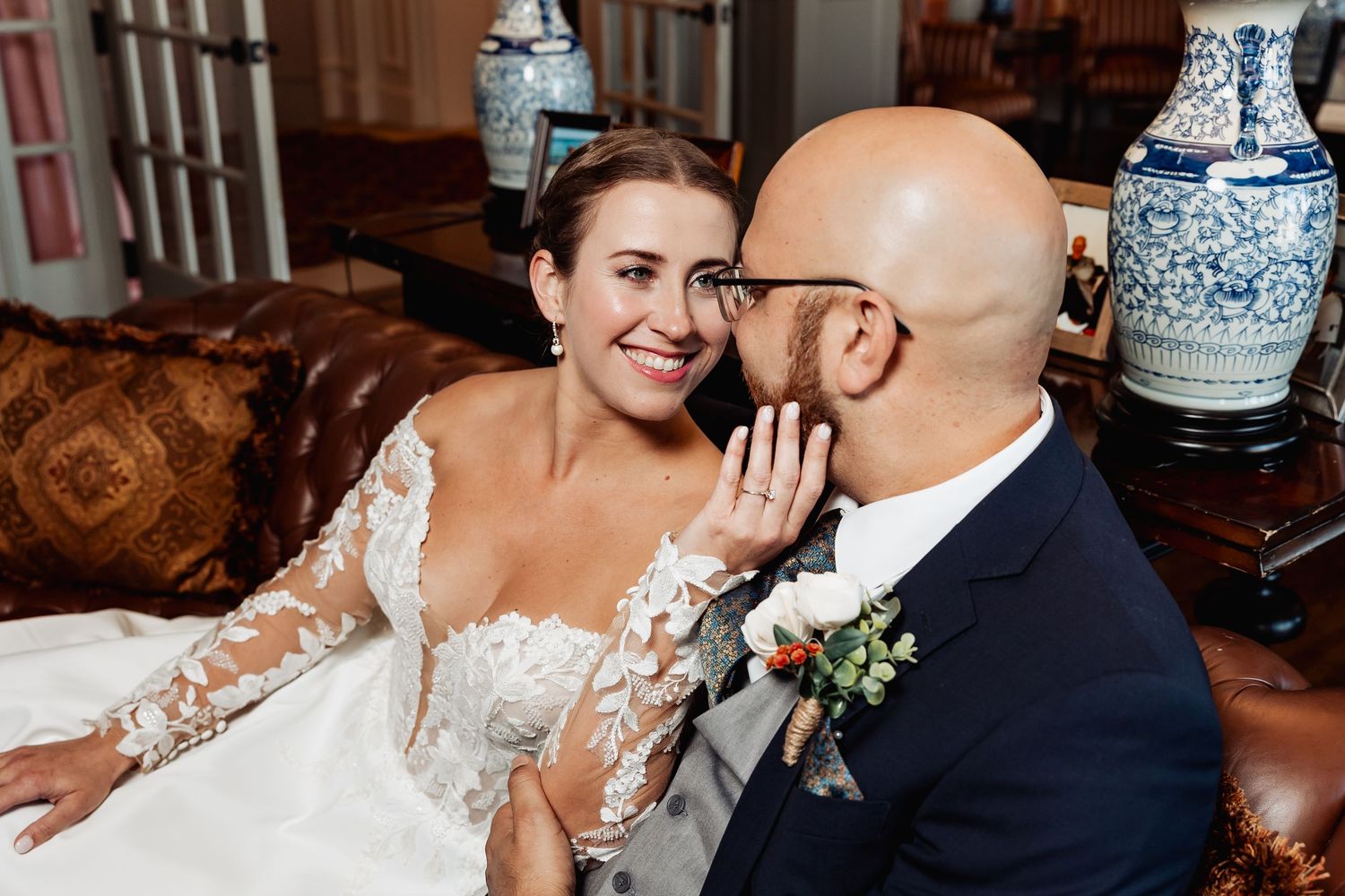 Smiling bride in lace gown and groom share a whispered secret on leather sofa at wedding.