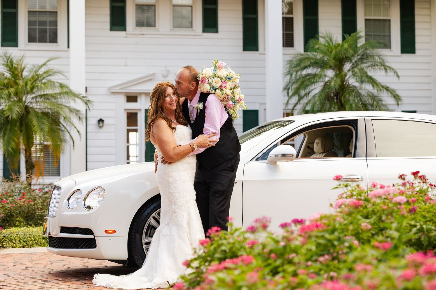 Bride and groom share a romantic moment beside a white Bentley luxury car at an elegant wedding venue.