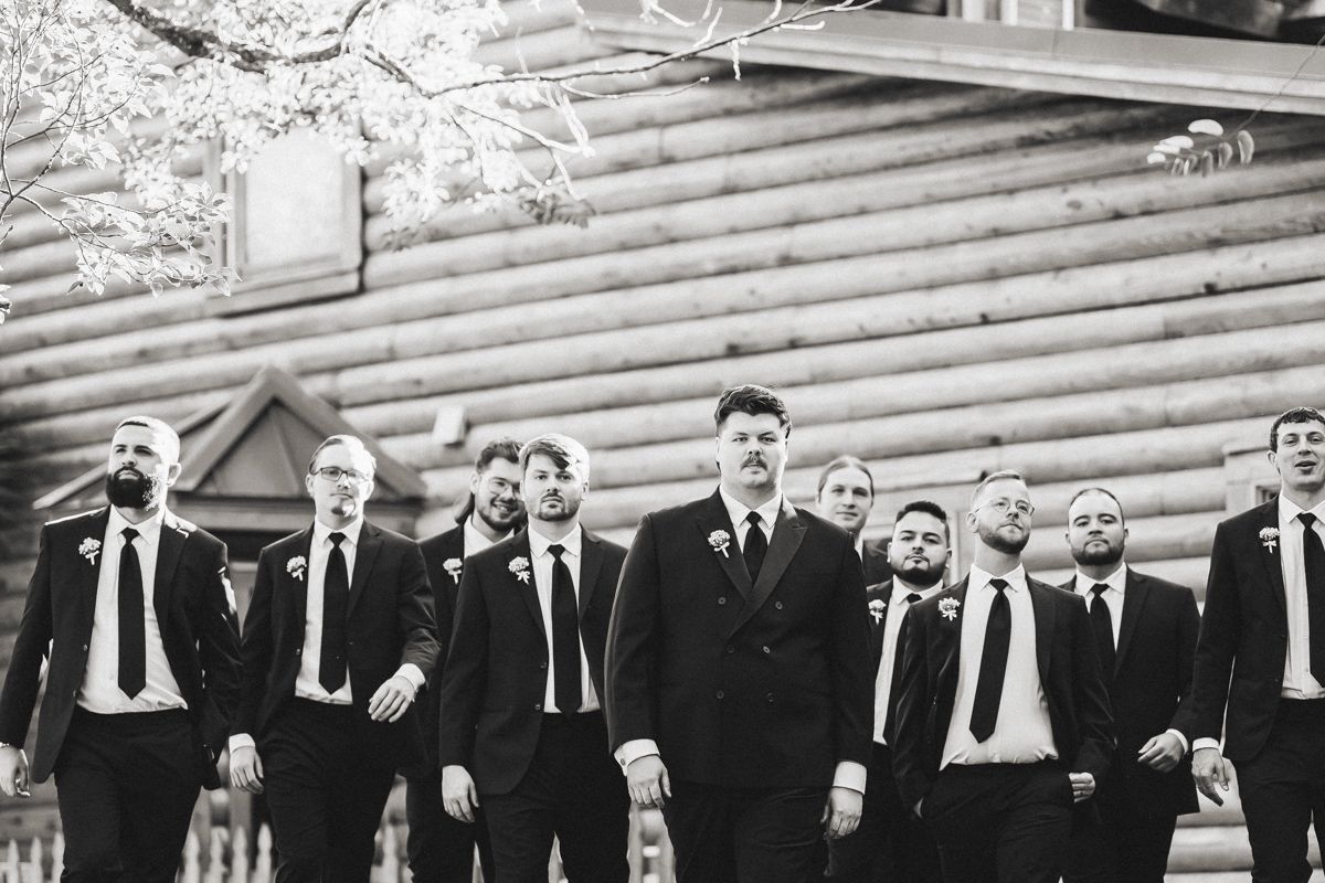 A group of groomsmen in black suits and ties stand together in a line outside a rustic log cabin.