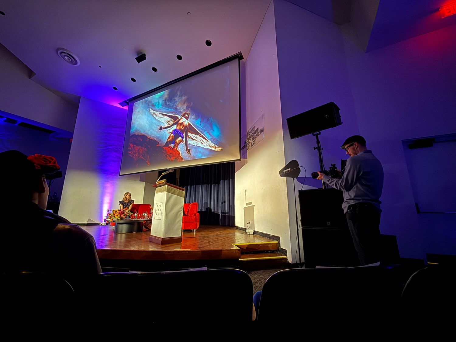 A panoramic view of a dimly lit room with blue accent lighting and projector screens showing presentation content.