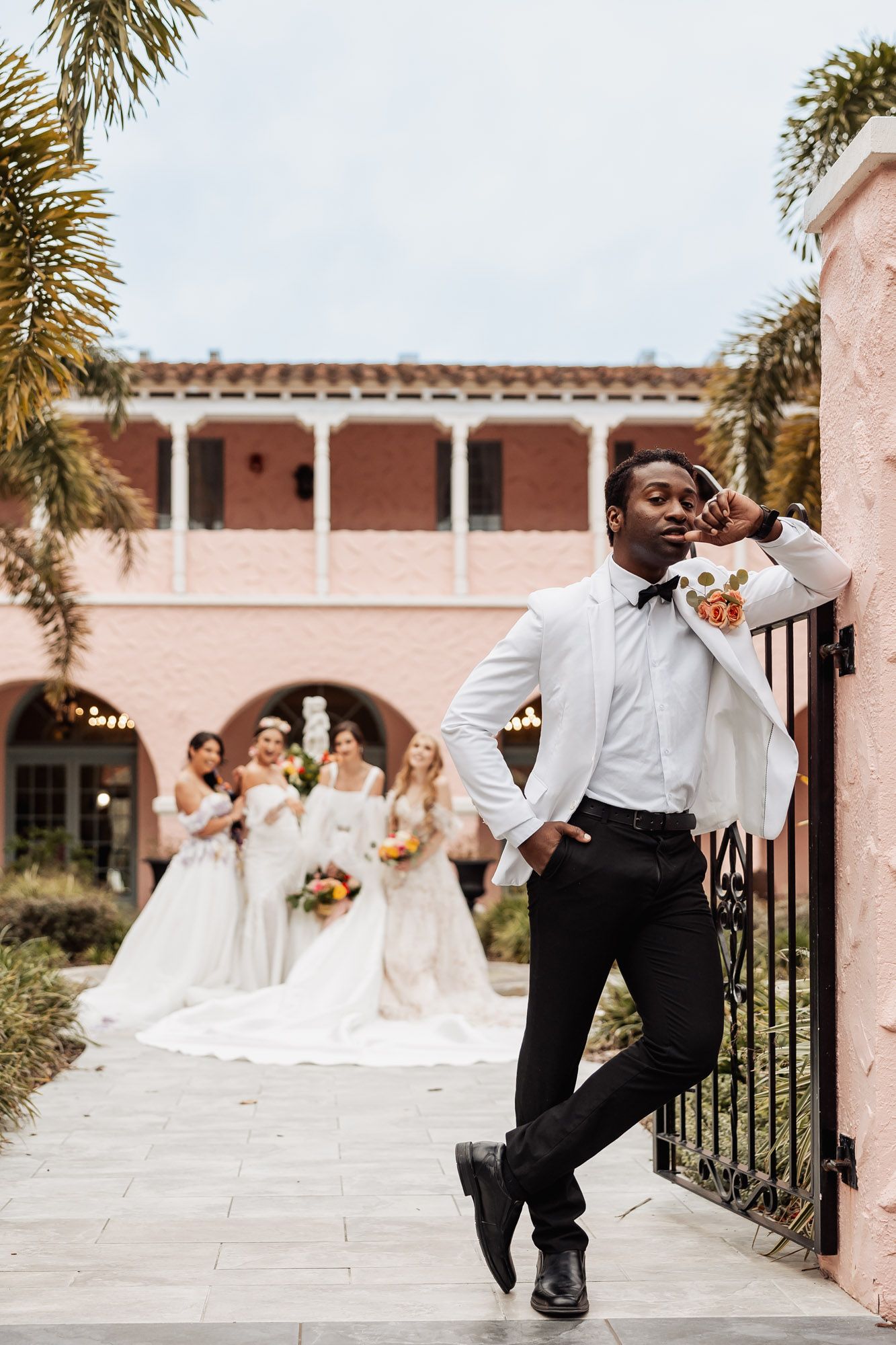 A member of the wedding party poses playfully at a pink Mediterranean-style venue with bridesmaids in white dresses in background.