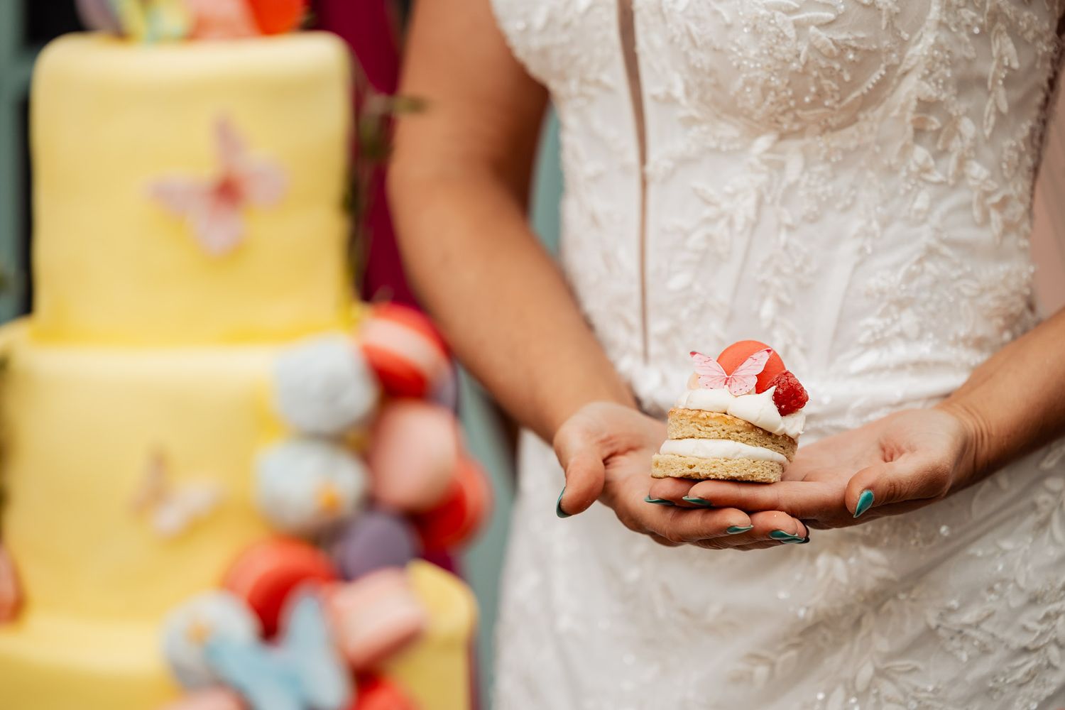 A delicate macaron with a strawberry garnish is held near an elegant yellow wedding cake decorated with flowers.