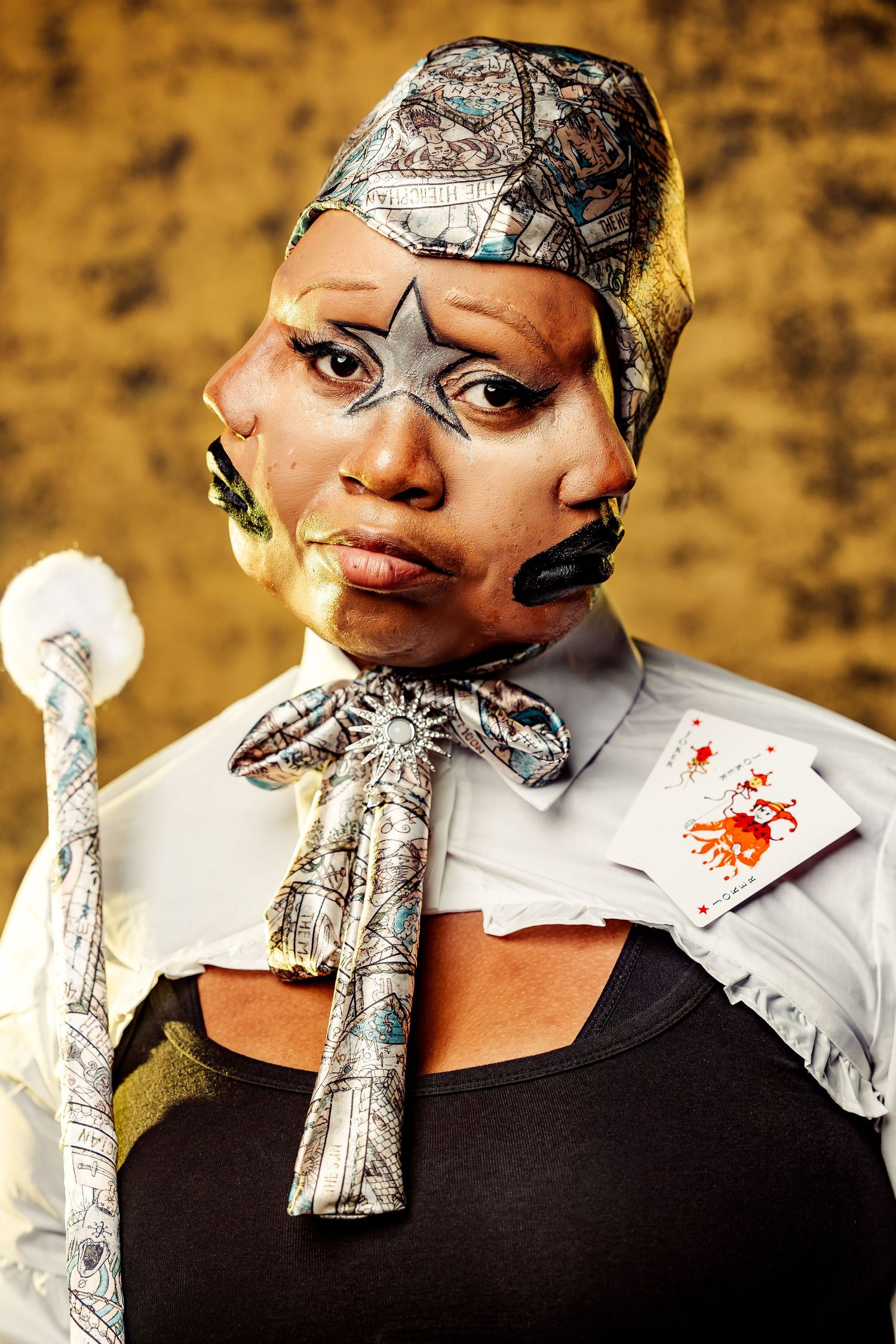 Portrait of performer in theatrical makeup wearing patterned headwrap and bow tie against gold background.
