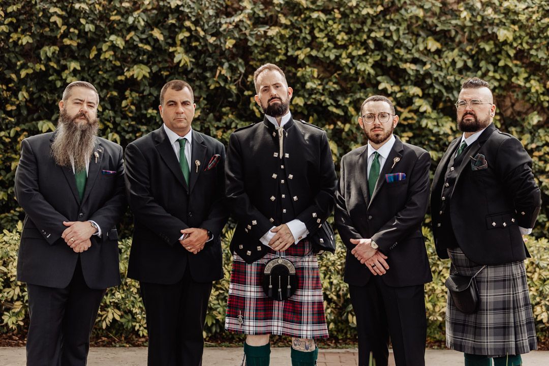 Five groomsmen pose outdoors, groom in center wearing tartan kilt and Scottish Highland dress.