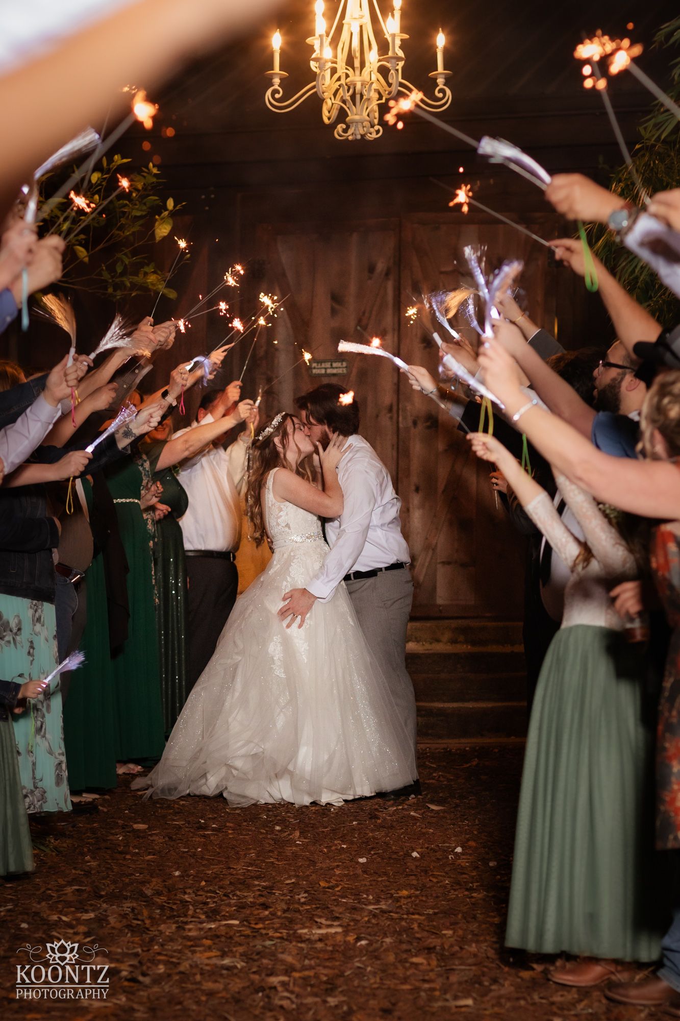 Bride and groom kiss during magical sparkler exit surrounded by cheering guests at barn wedding.