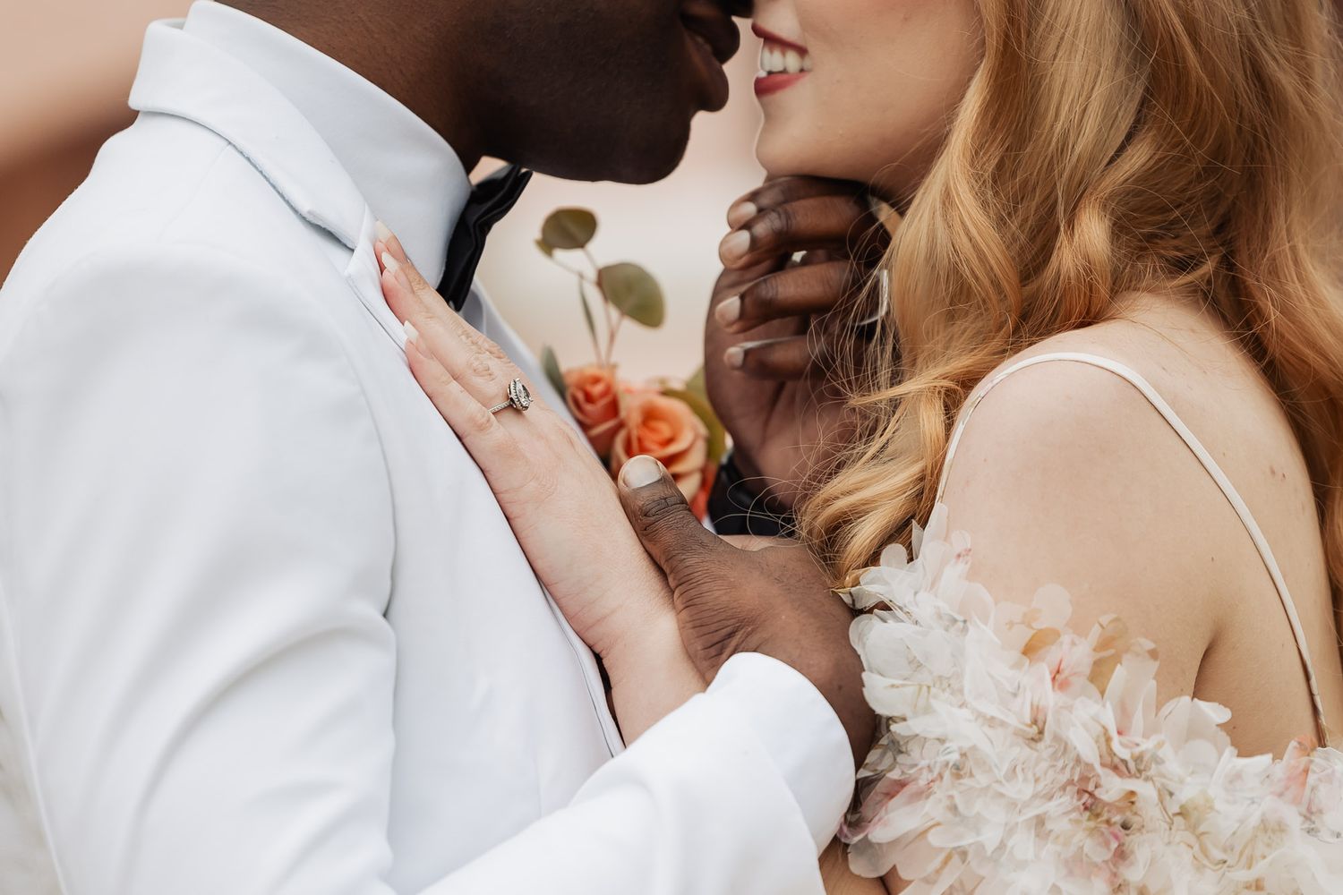 Close-up of interracial wedding couple nearly kissing, bride in floral gown with diamond ring visible.