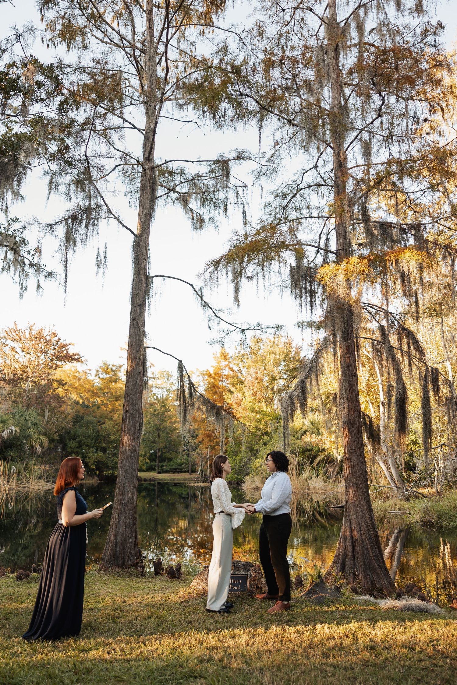 Two women exchanging vows in an outdoor handfasting ceremony beside a pond with cypress trees.