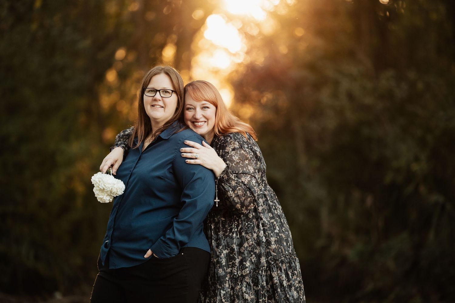 Two women embrace warmly in a sun-drenched forest setting, smiling together during a romantic engagement photoshoot.