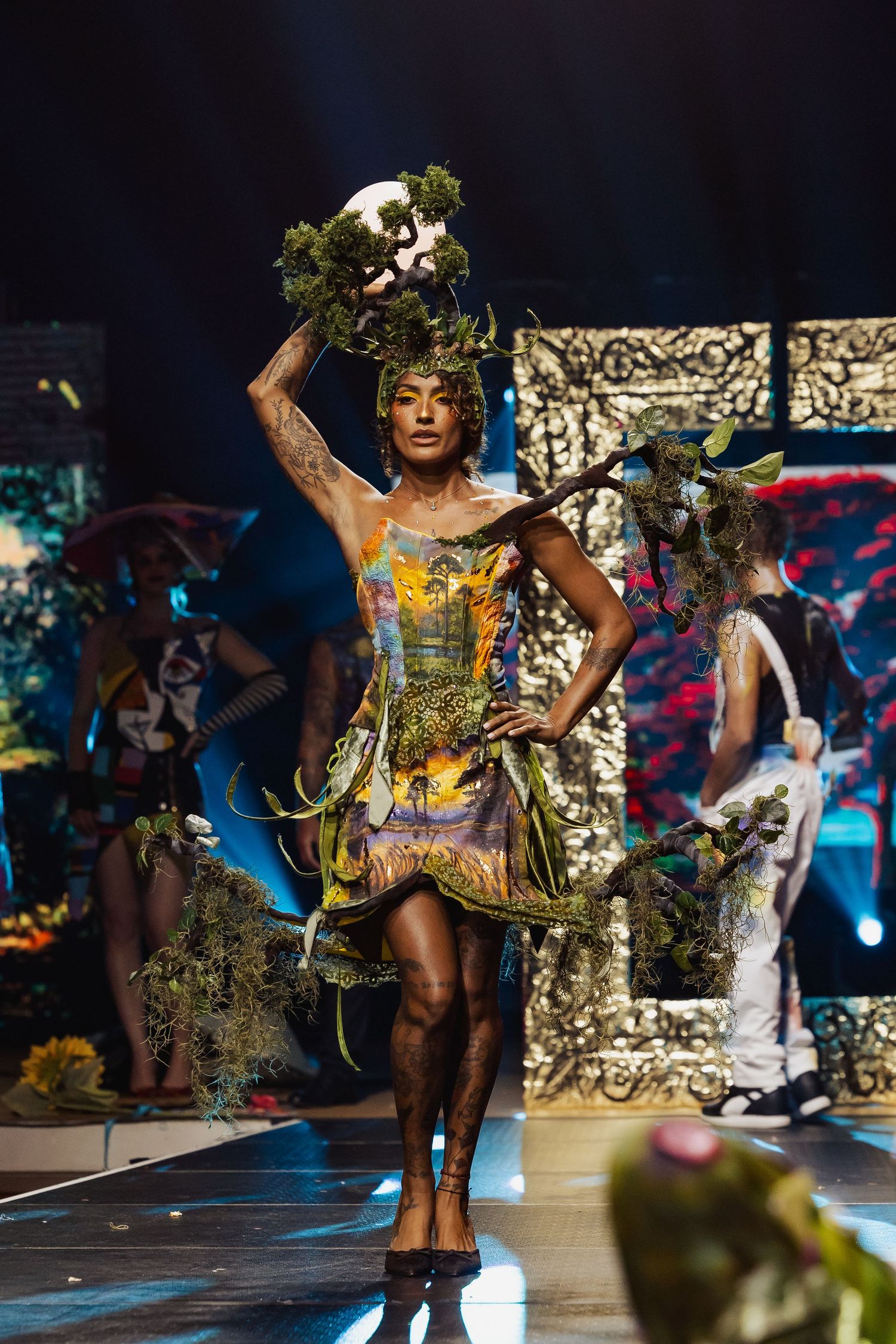 Model in nature-inspired dress holding plants and skull headpiece on runway.