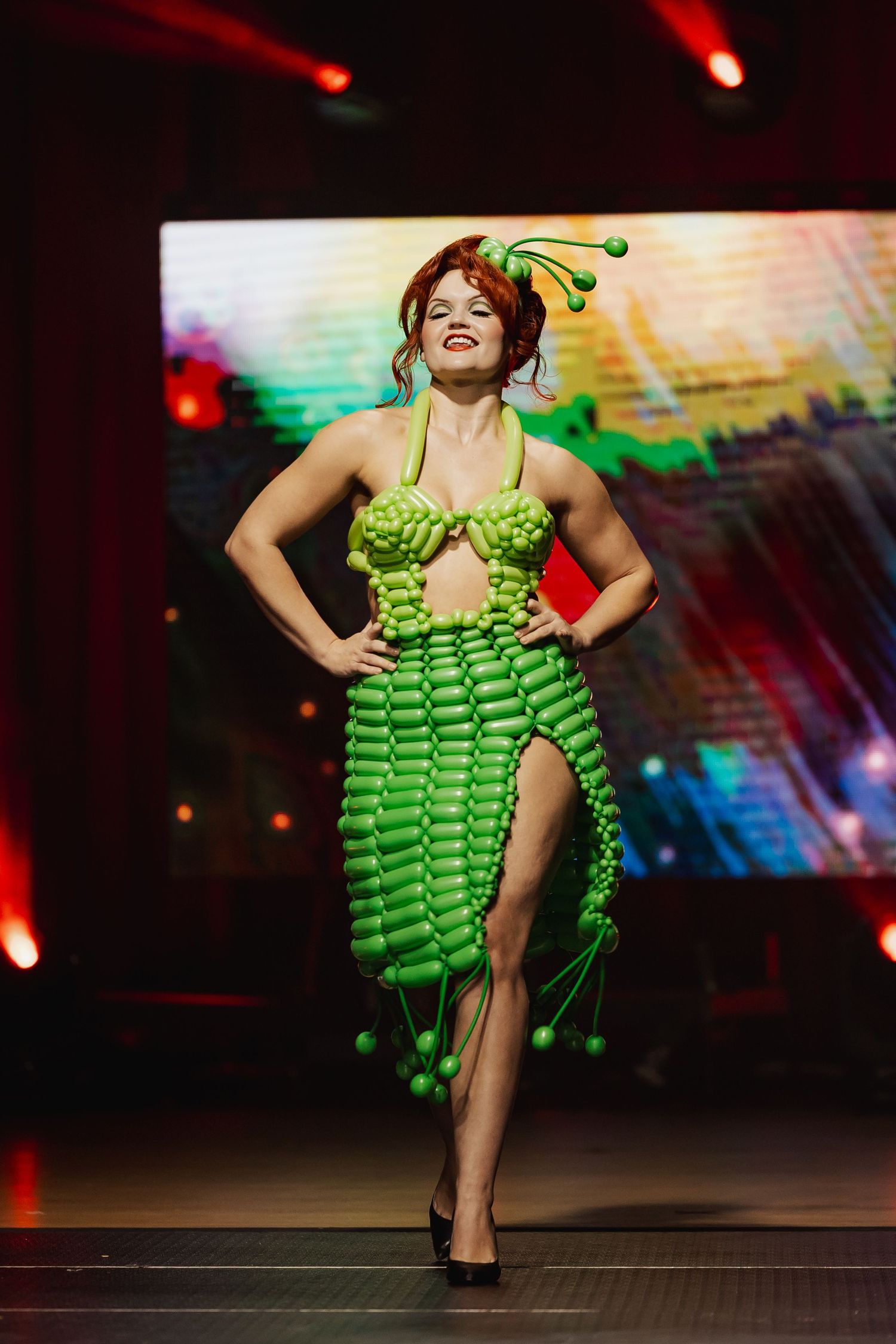 Female model walks runway in green balloon halter dress with matching sculptural headpiece.