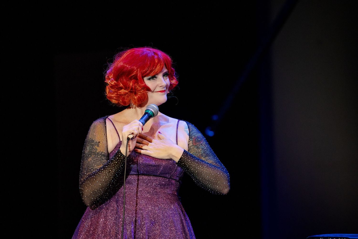 Red-haired female singer in purple glitter dress performing on stage with hand on chest.