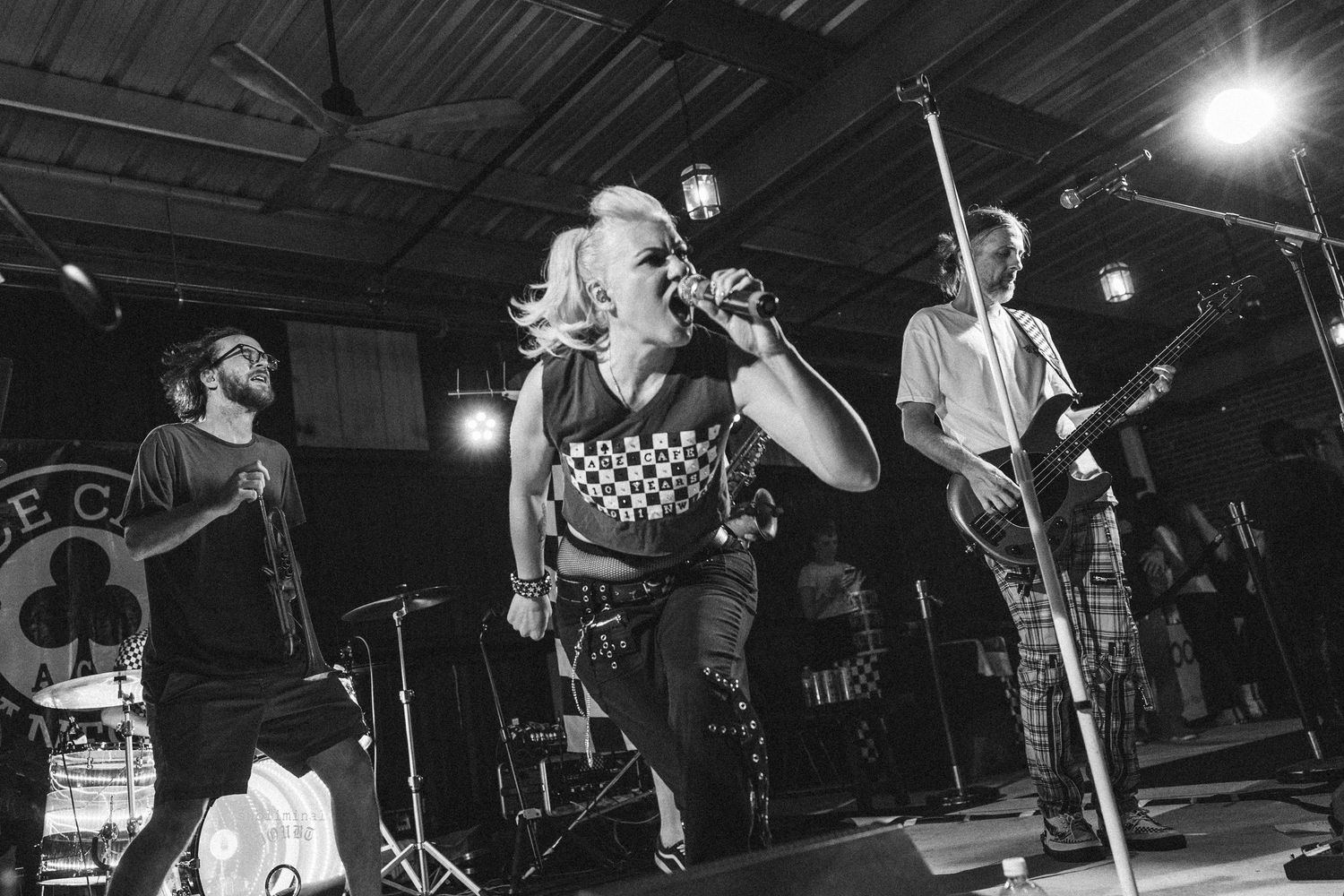 Female punk vocalist performs energetically on stage with band in black and white photo.