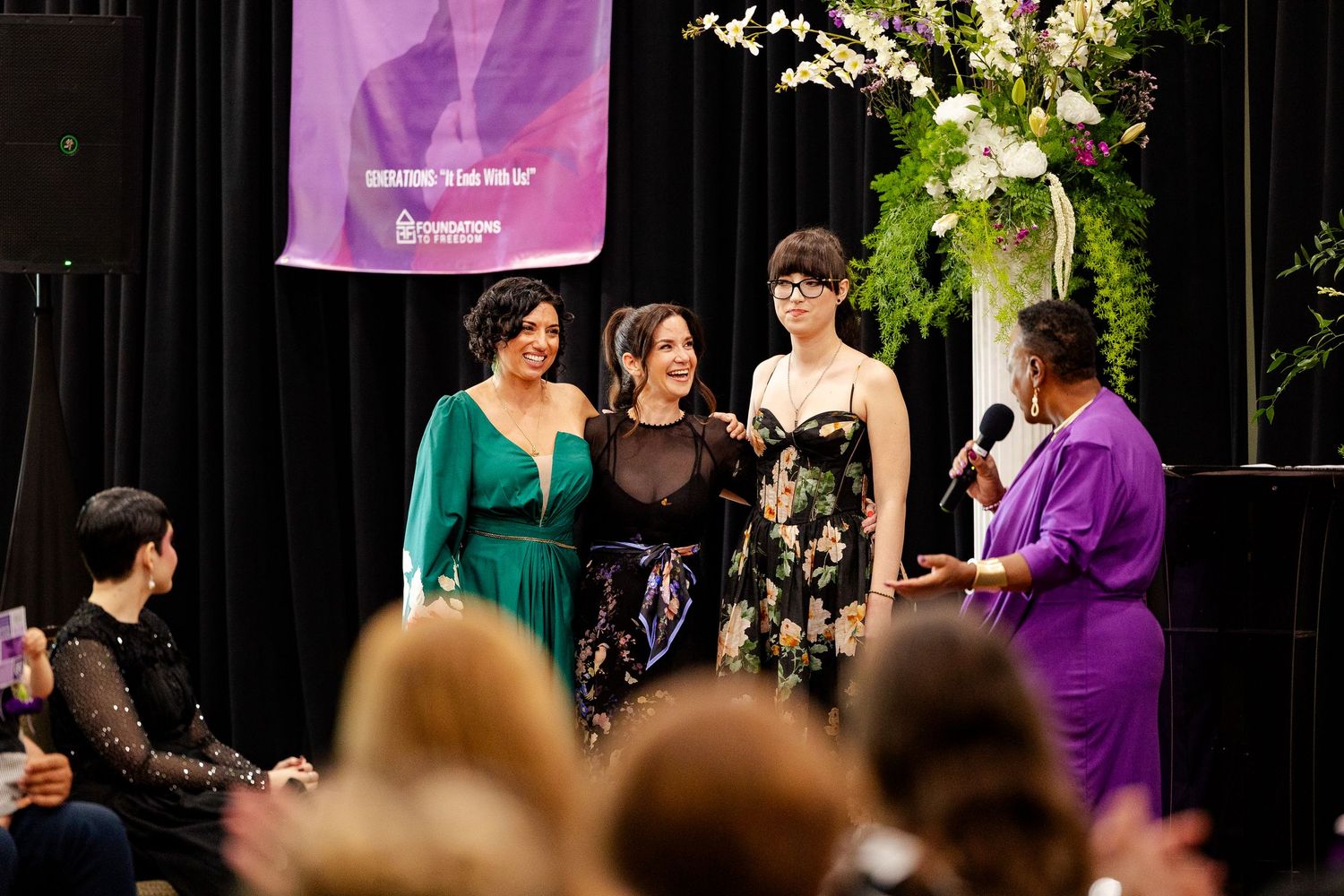 Four women stand together on stage being interviewed at elegant fashion event.