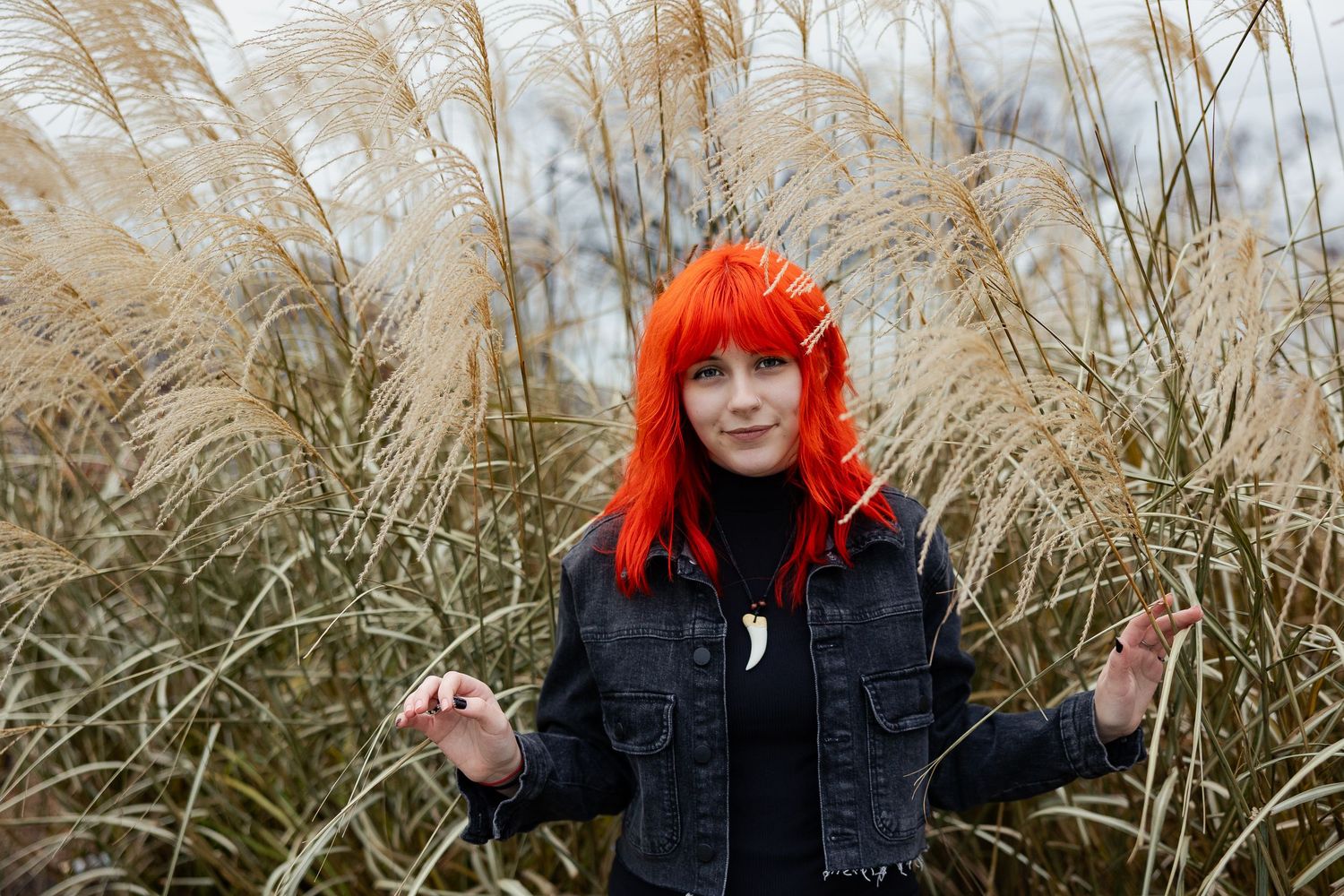 Young woman with vibrant orange hair posing outdoors among tall golden grass in a black denim jacket.