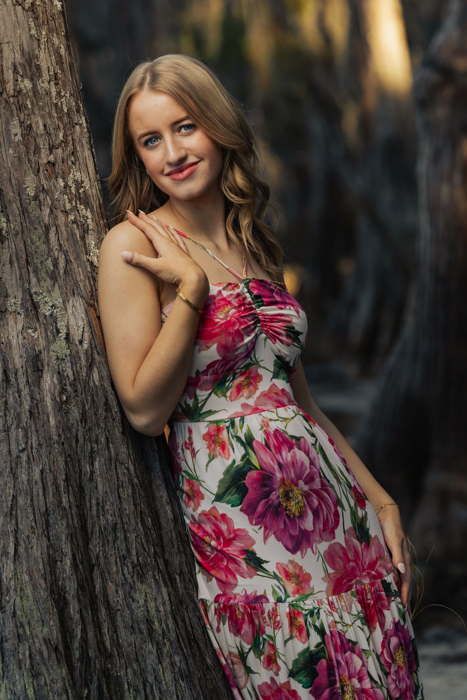 Young woman in floral dress leaning against tree, smiling outdoors in warm golden light.
