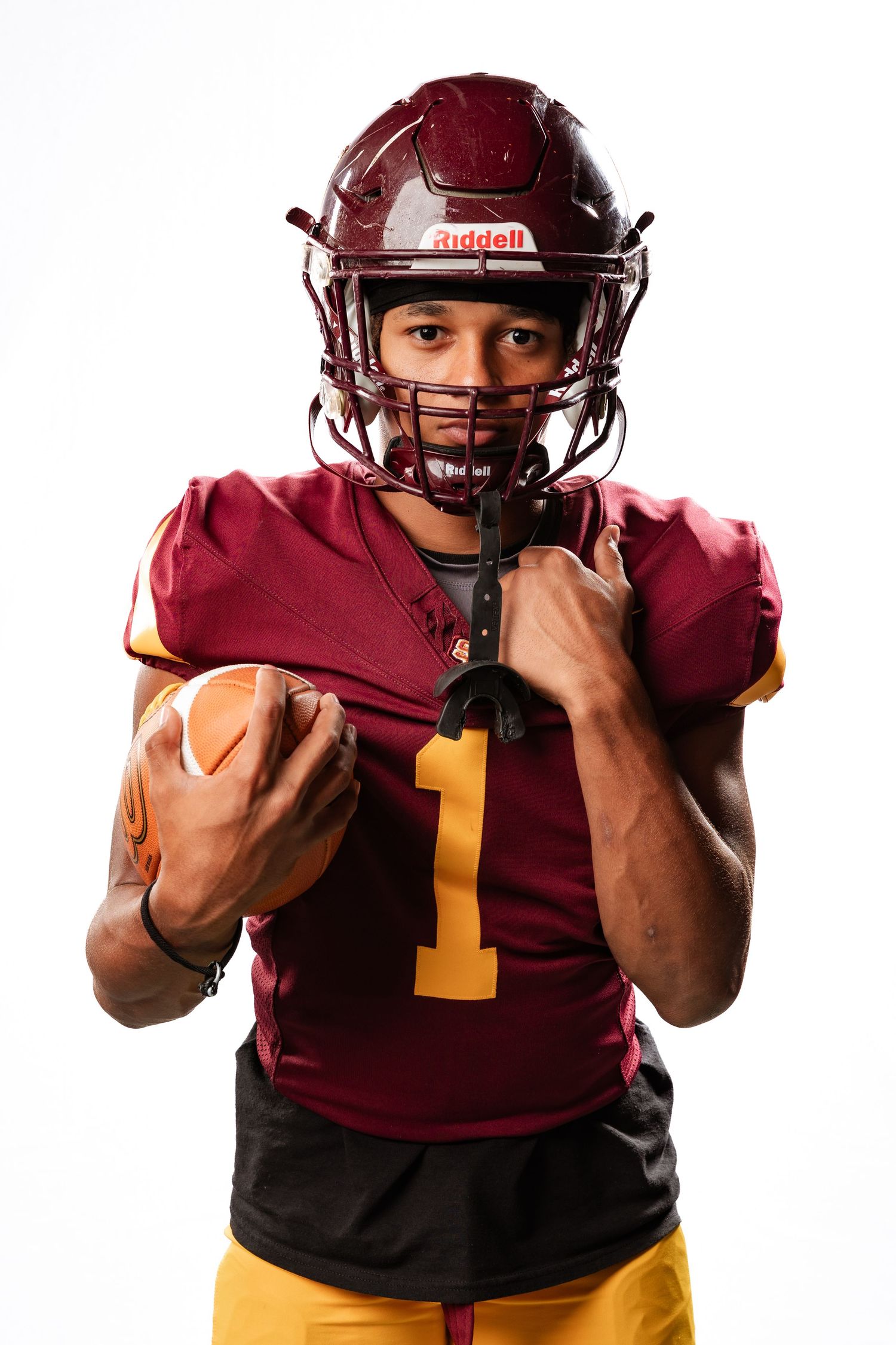 American football player in maroon Riddell helmet and jersey number 1 holding football against white background.
