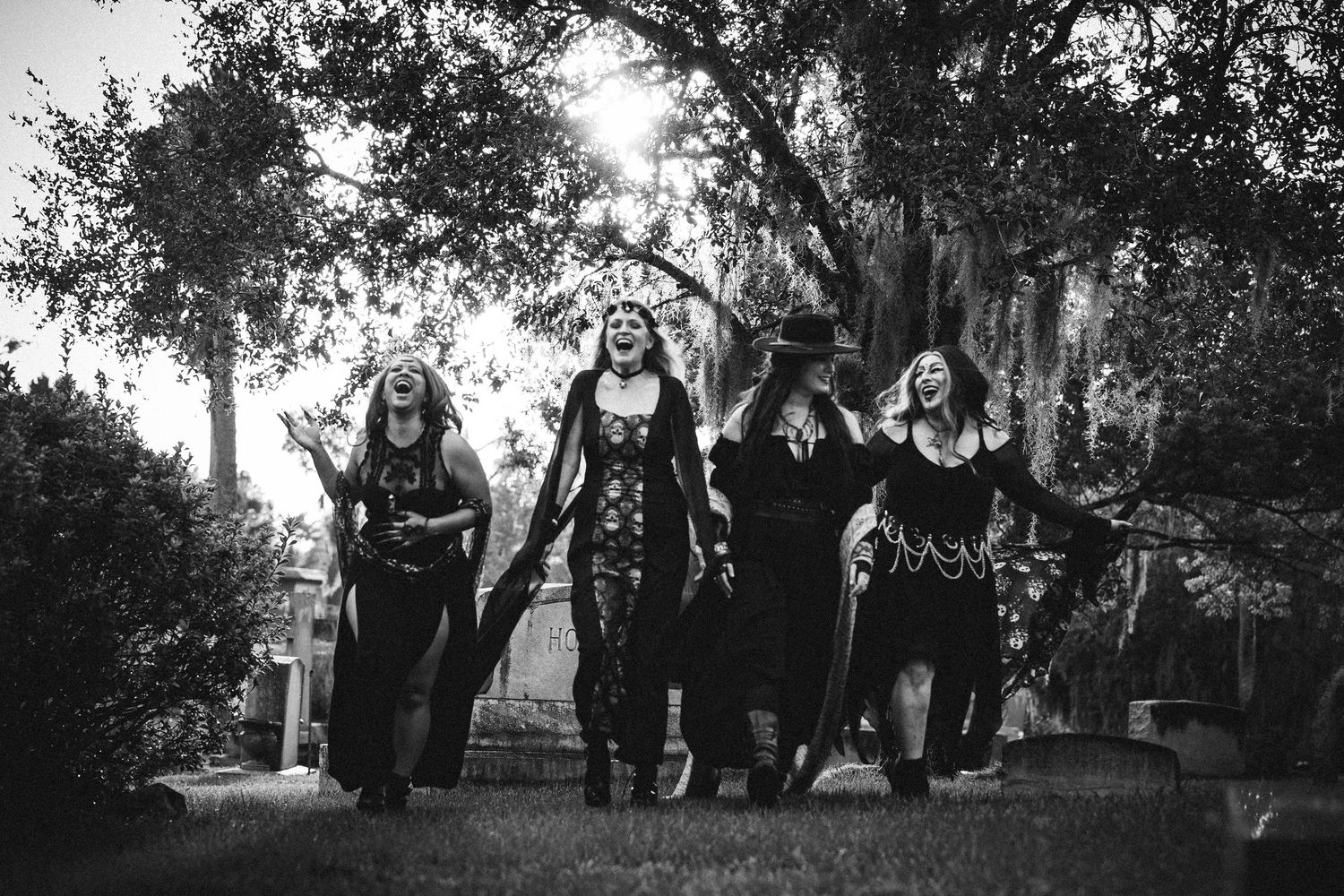 Black and white photo of four women in witch costumes walking together through a garden setting.