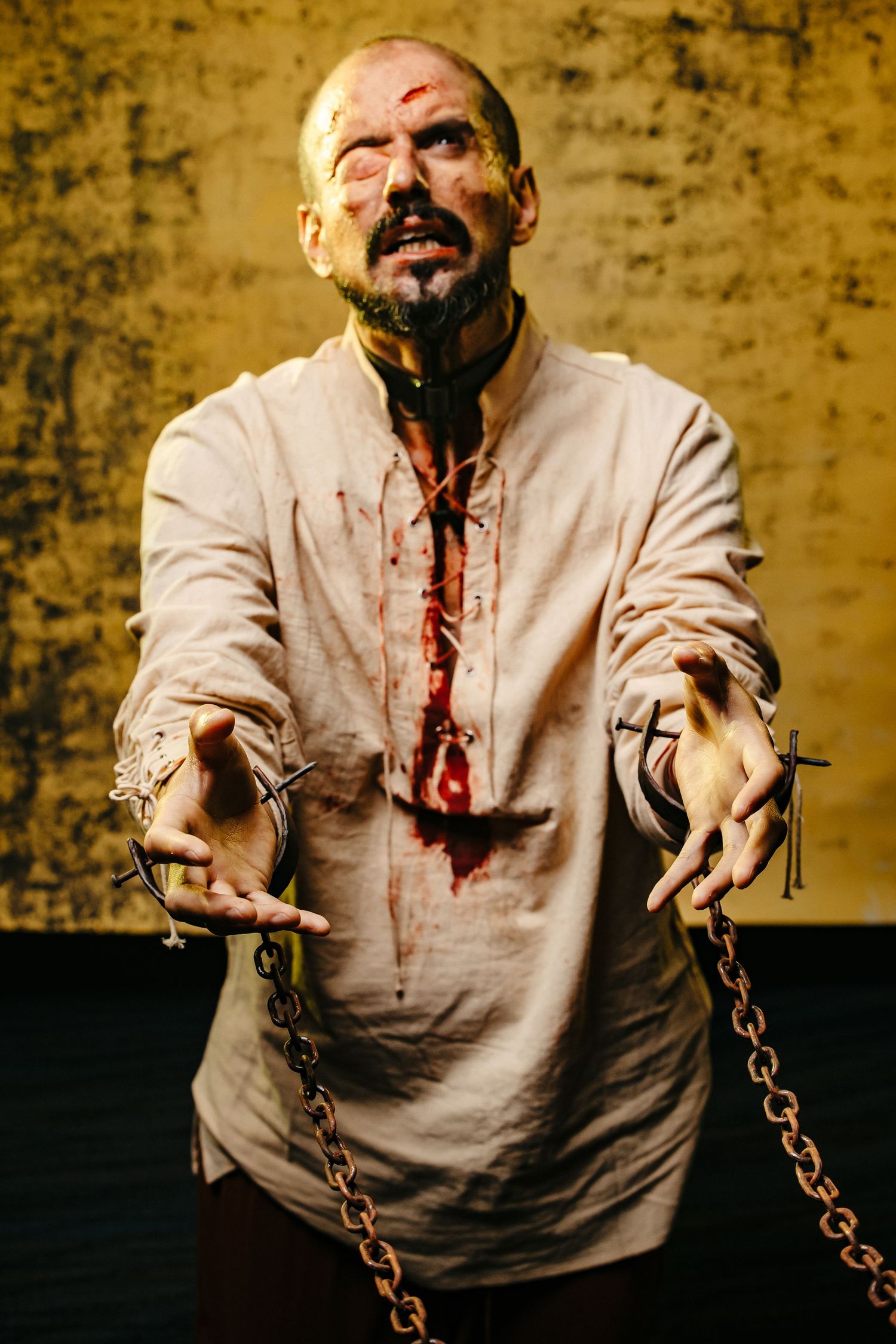 Bloodied man in tattered shirt extends chained hands forward in a dramatic Halloween or theatrical scene.