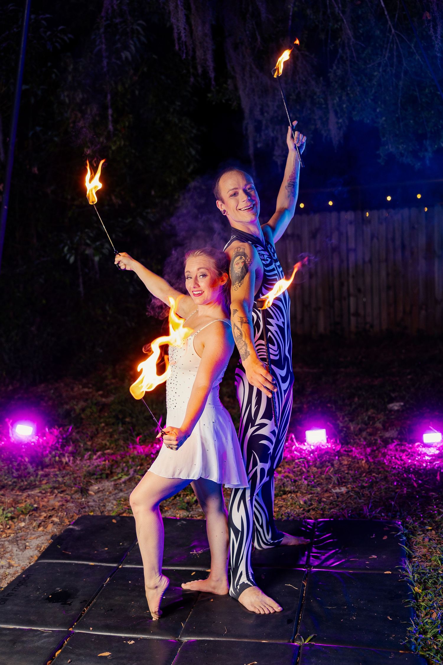 Two barefoot fire performers stand back-to-back at night holding flaming torches amid colorful purple stage lighting.