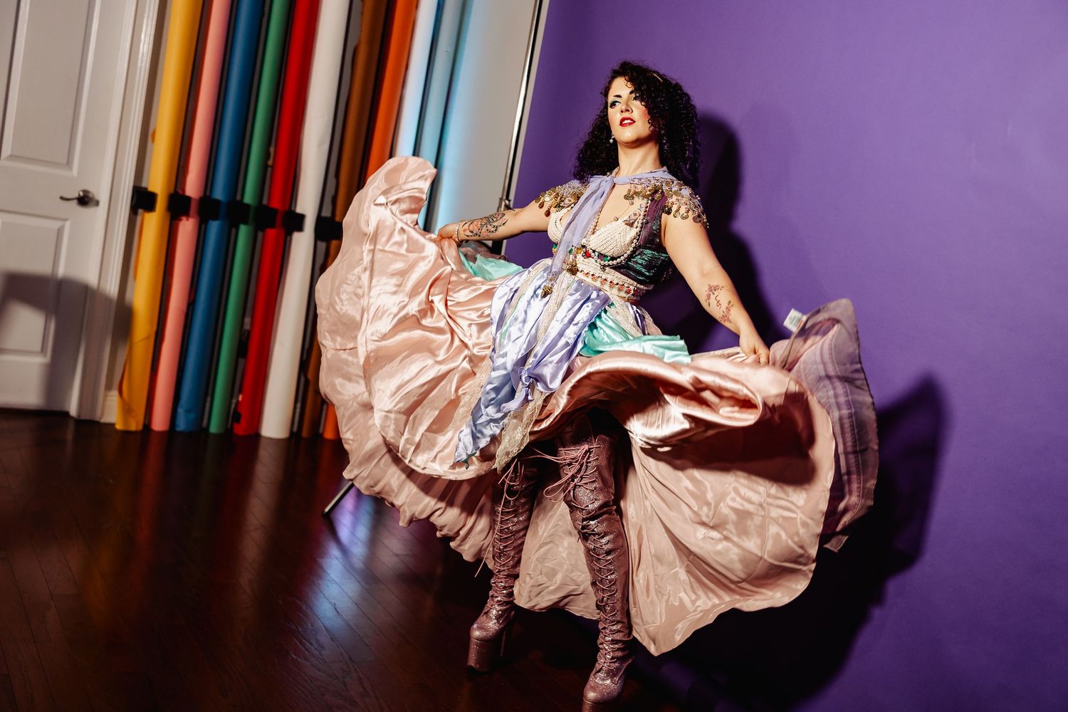 Woman in swirling pastel belly dance costume twirls on hardwood floor near colorful fabric rolls.