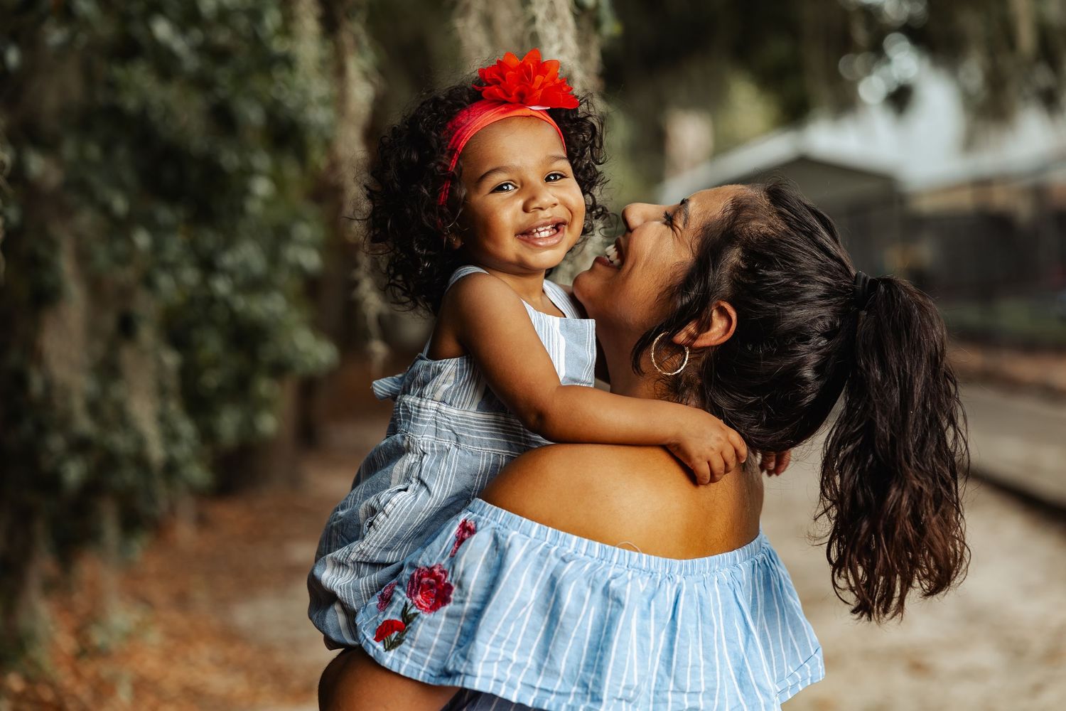 A happy toddler girl with a red flower headband hugs her mother outdoors.