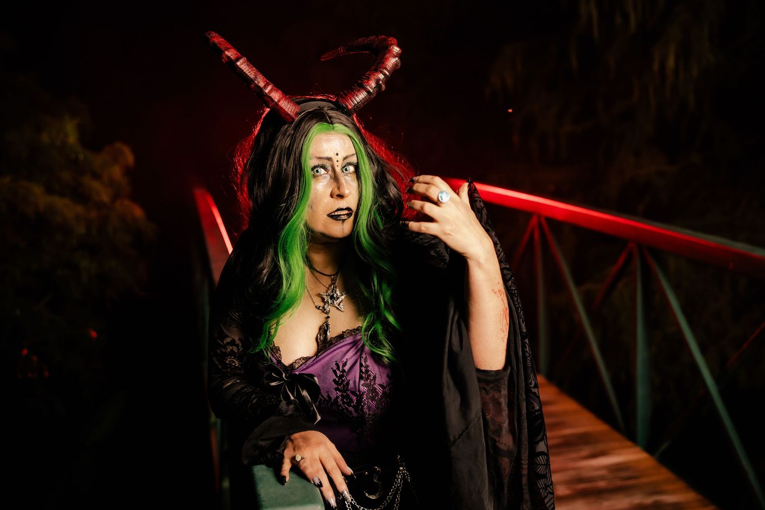 Woman with green hair and red demon horns in gothic costume poses on a bridge with red lighting at night.