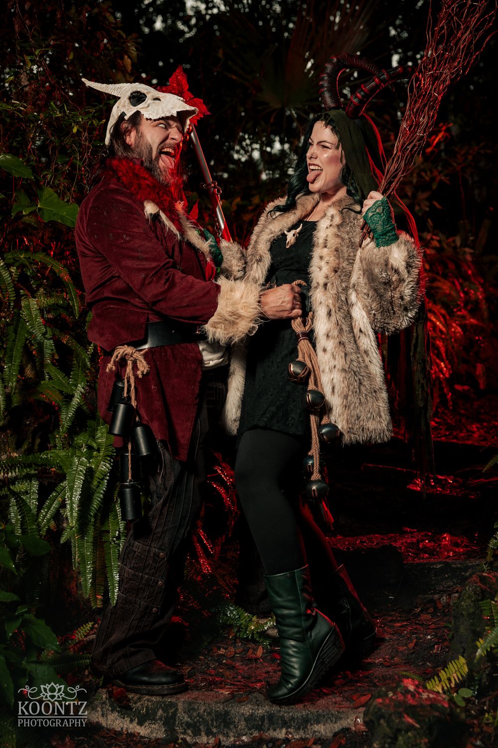 Two people in dark fantasy Halloween costumes laugh together in a moody forest setting by Koontz Photography.